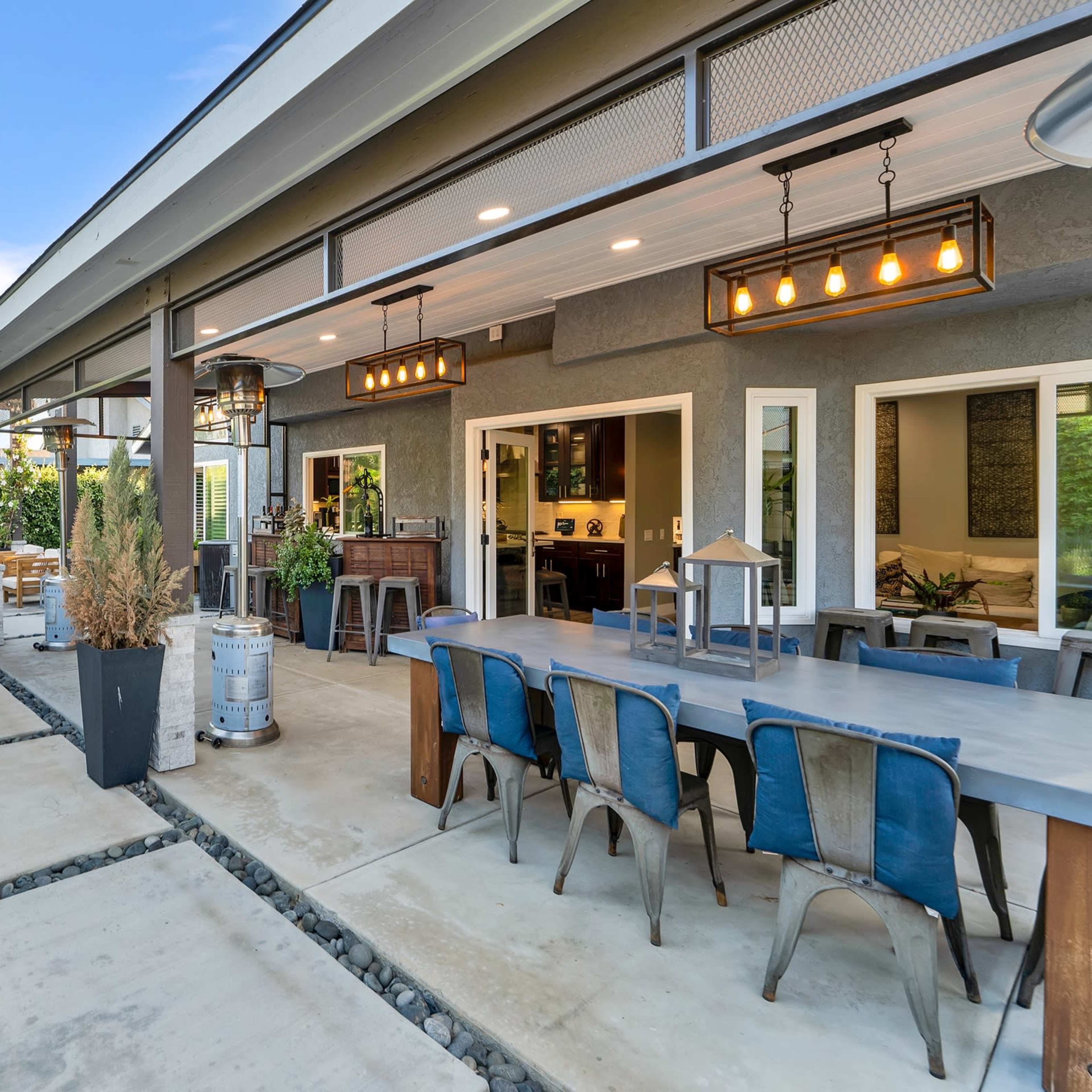 A modern outdoor patio area with a dining table, lounge seating, and a bar, surrounded by lush greenery and a stone pathway.