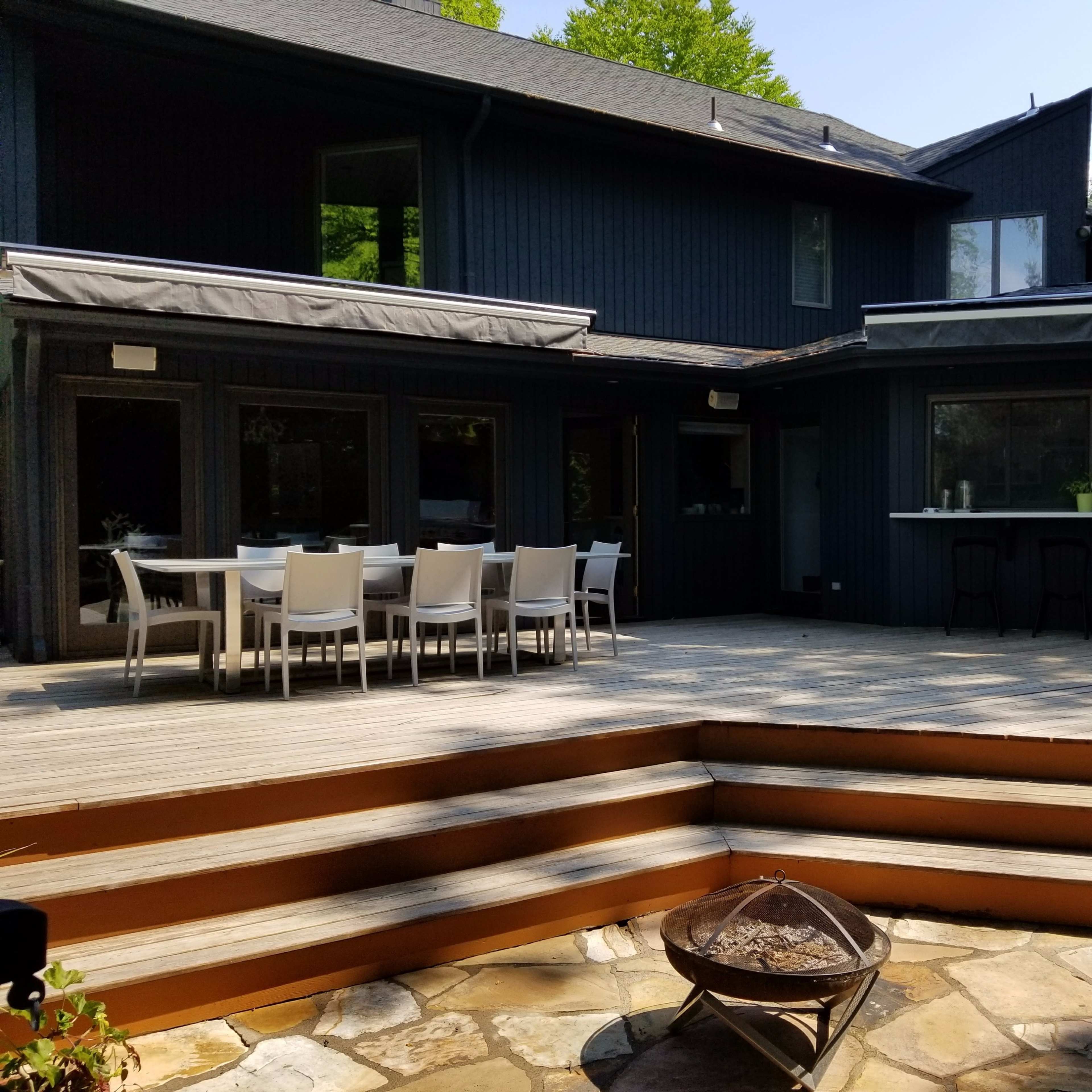 A spacious wooden deck with a dining area and a barbecue setup is connected to a dark-colored house surrounded by greenery.