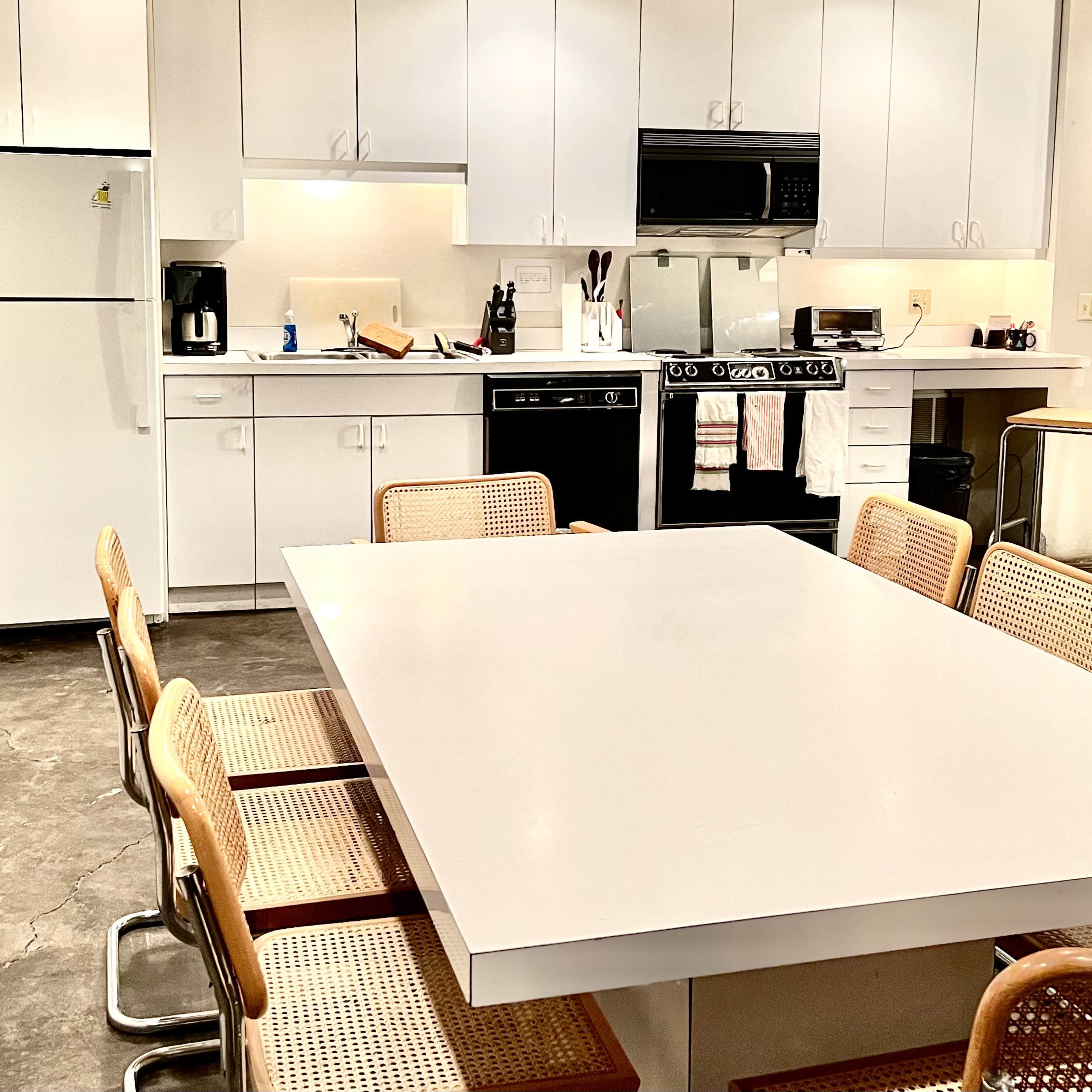 A modern kitchen features a large white table surrounded by eight cane-seat chairs, with appliances and cabinets in the background.