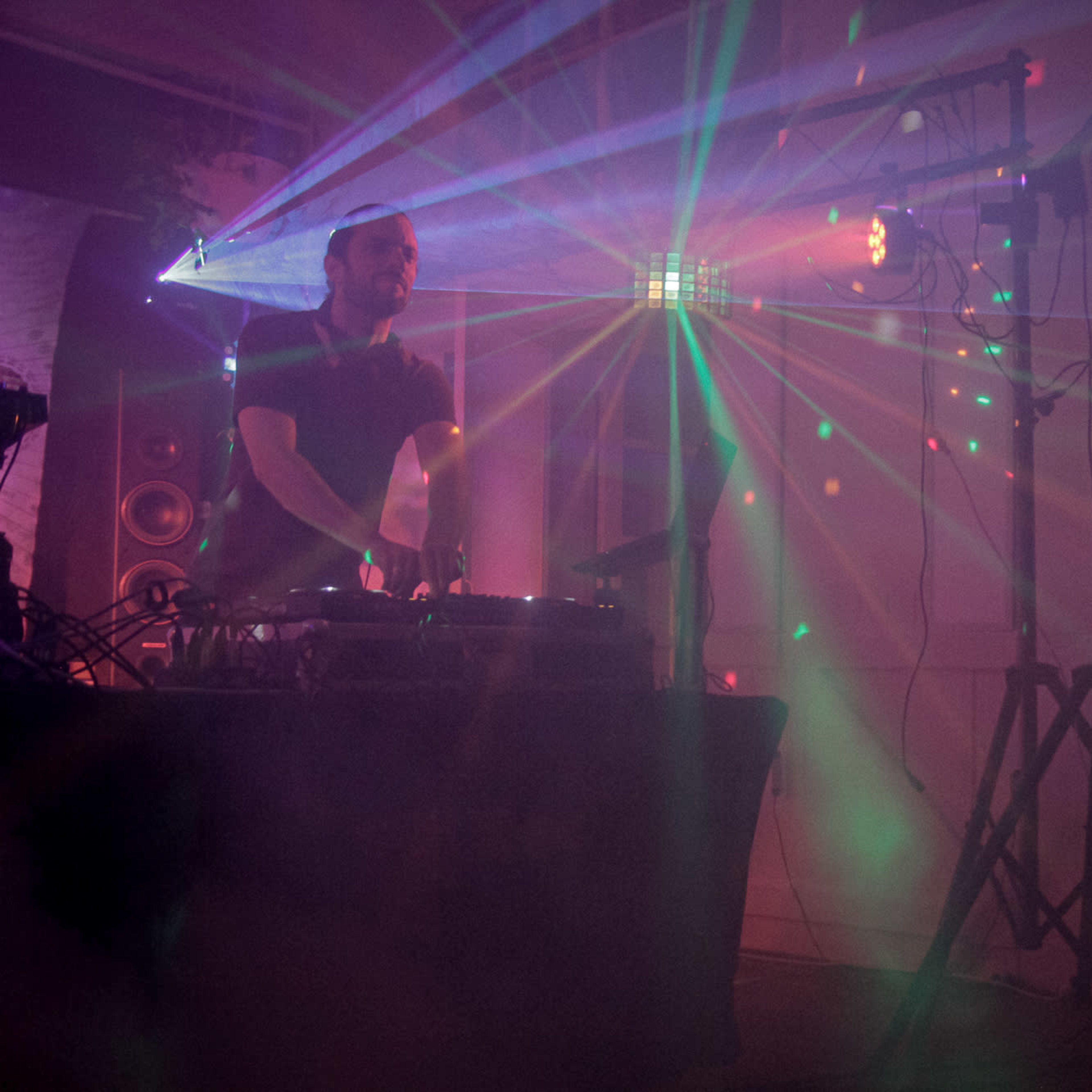 A DJ stands behind a mixer as colorful laser lights project beams and patterns onto the walls of a dimly lit room.
