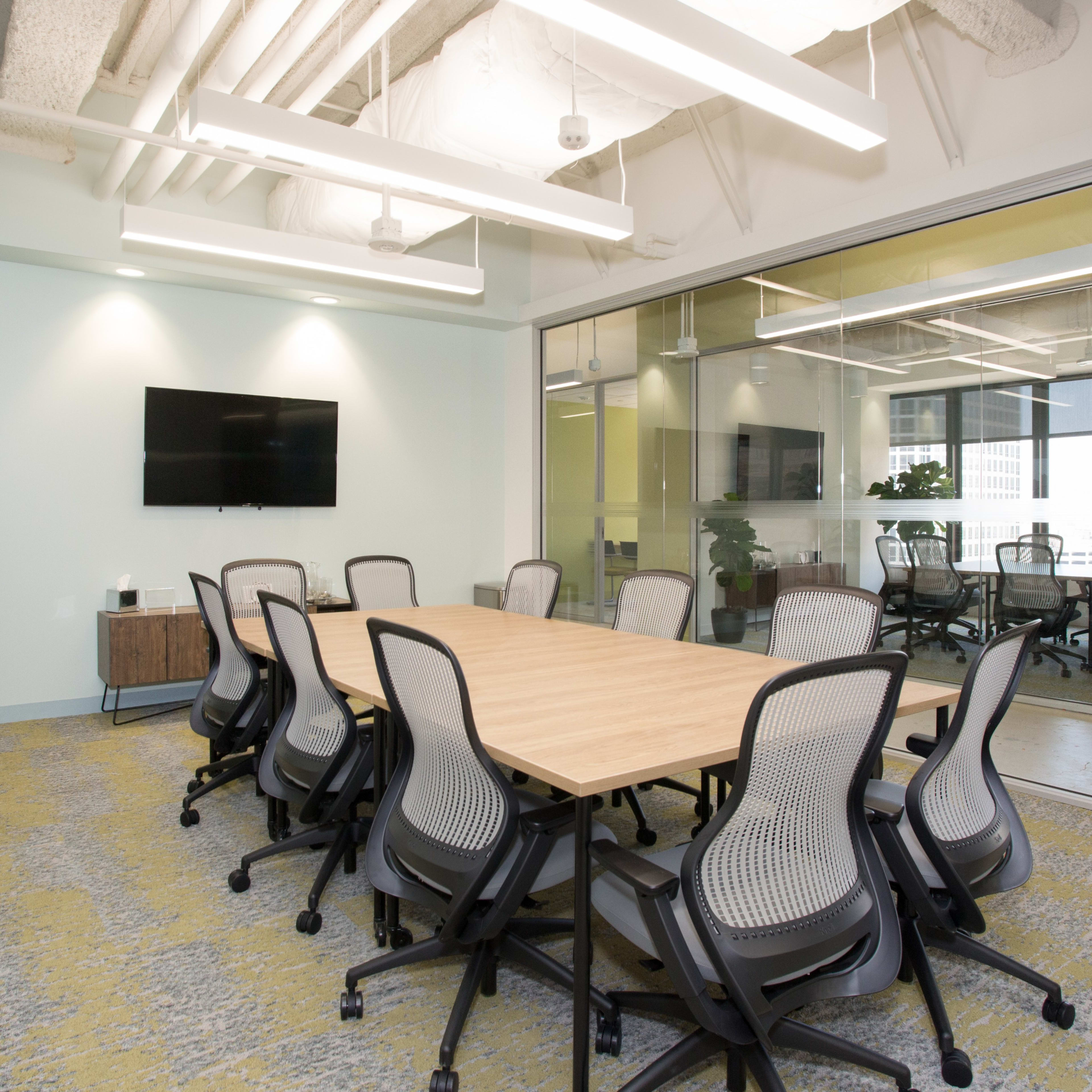 A modern conference room features a large wooden table surrounded by ergonomic chairs, with glass walls and a wall-mounted TV.