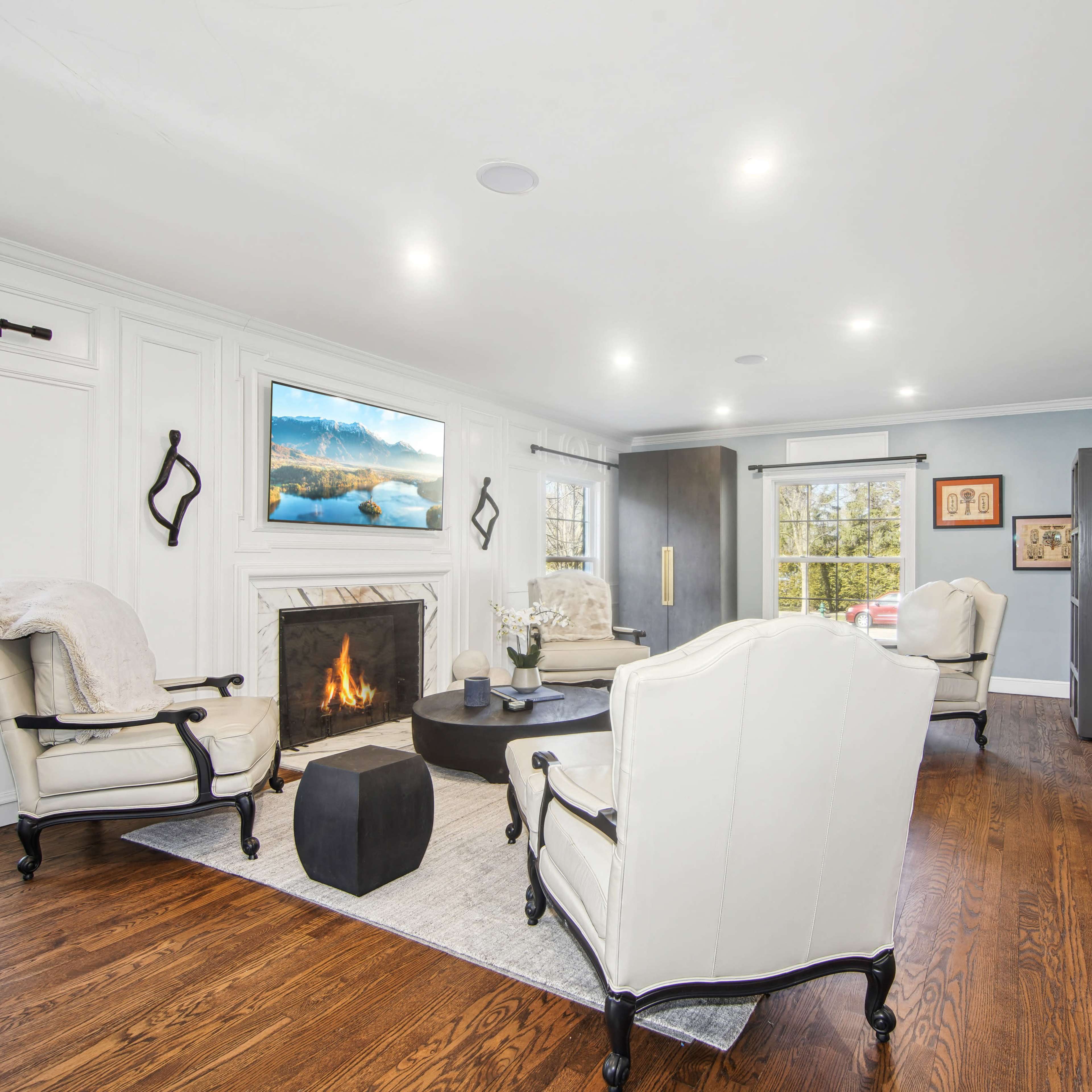 A modern living room features two white armchairs, a black coffee table, a fireplace, and a mounted television, with large windows allowing natural light in.