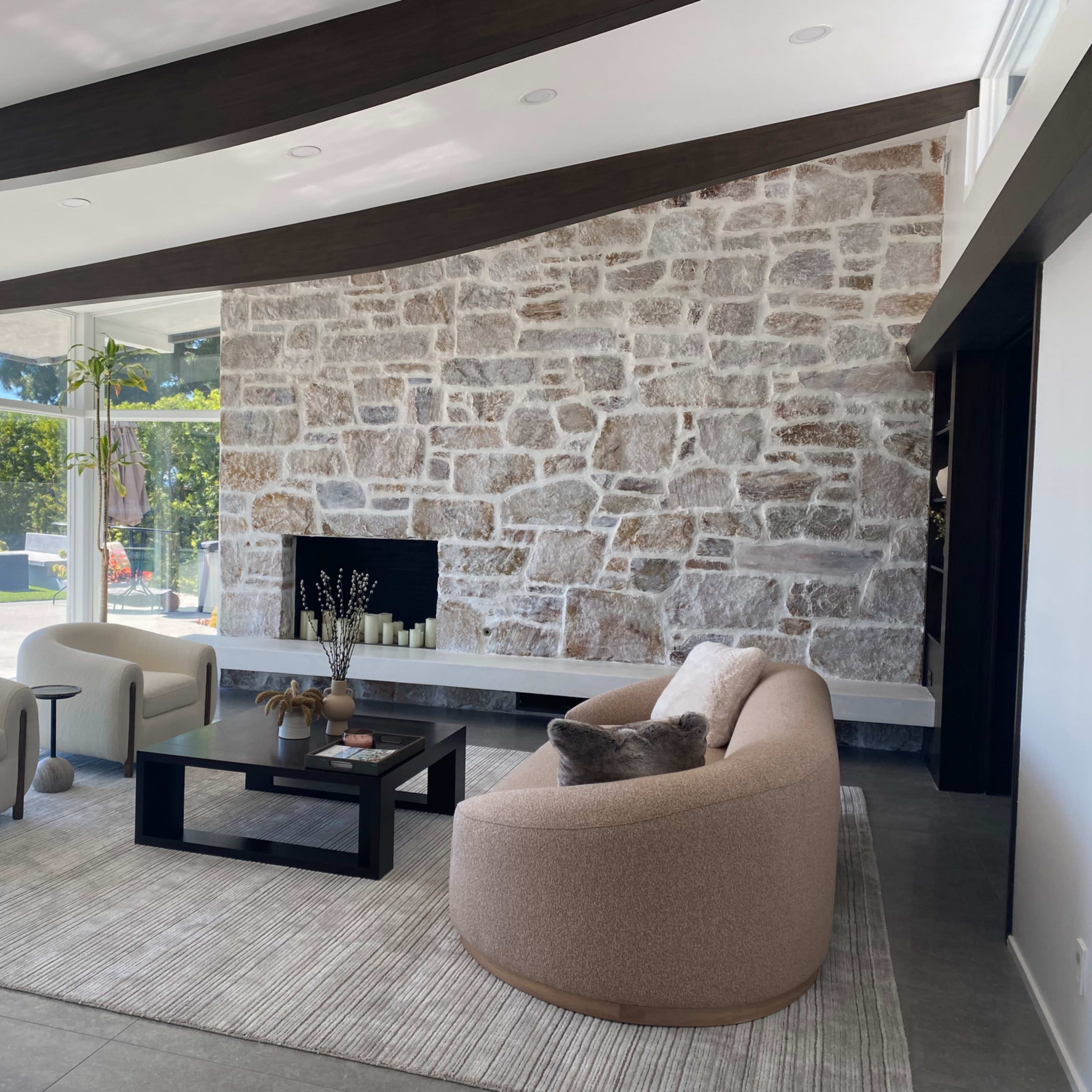 A modern living room with a stone accent wall, two curved sofas, a coffee table, and large windows providing natural light.