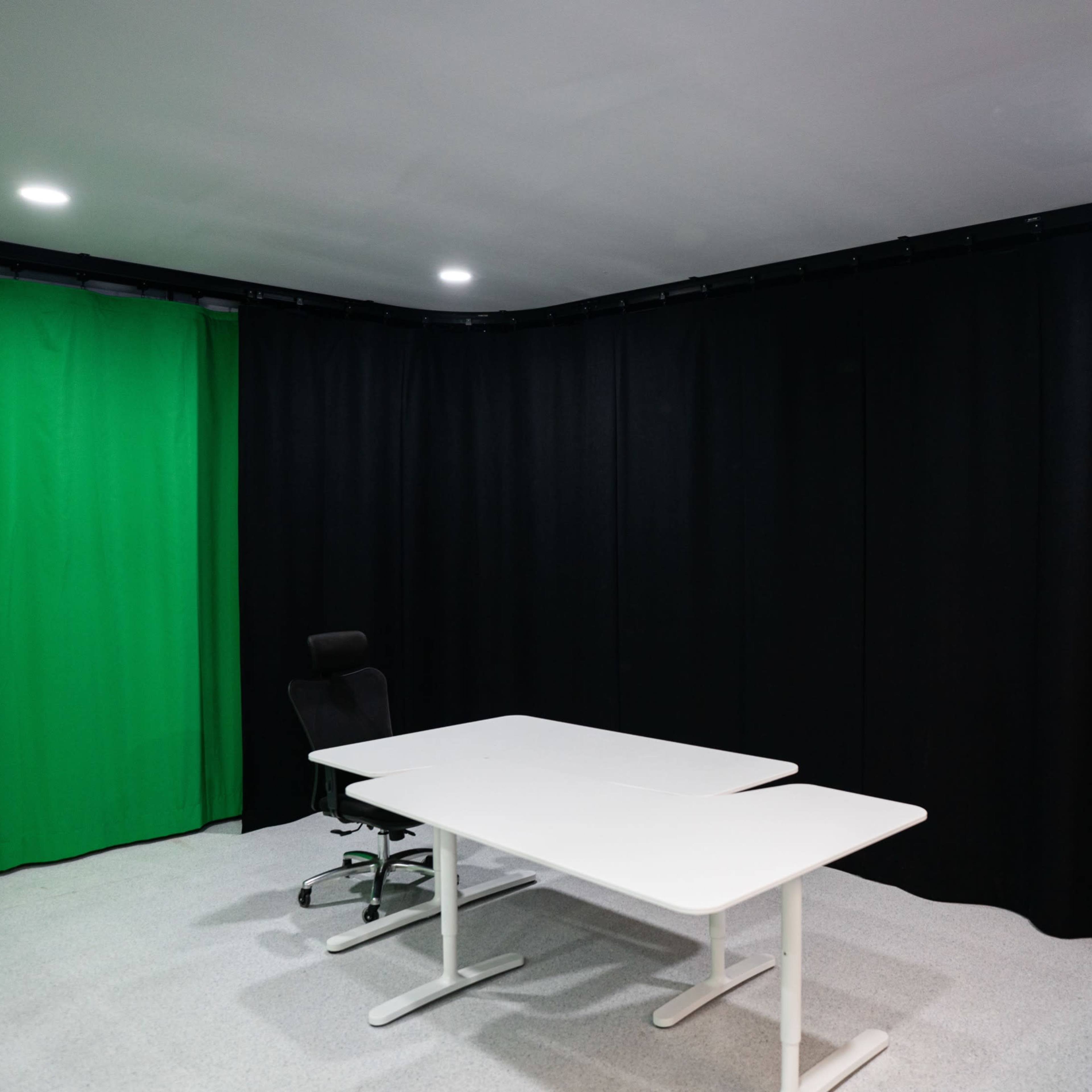 A small studio room features a green screen backdrop, black curtains, a white table, and an office chair.
