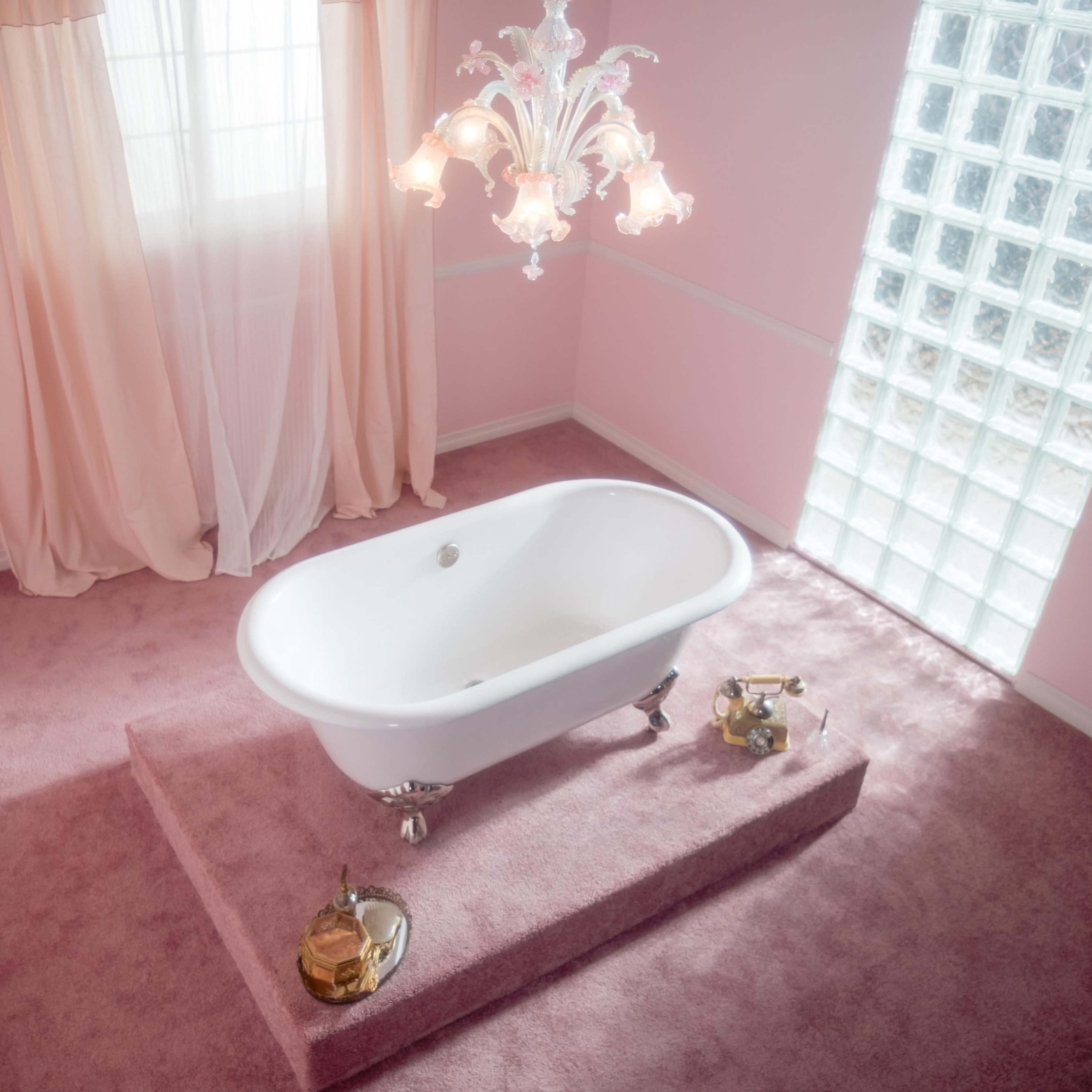 A white freestanding bathtub is positioned on a raised platform in a softly lit pink bathroom with a chandelier and a window featuring glass blocks.