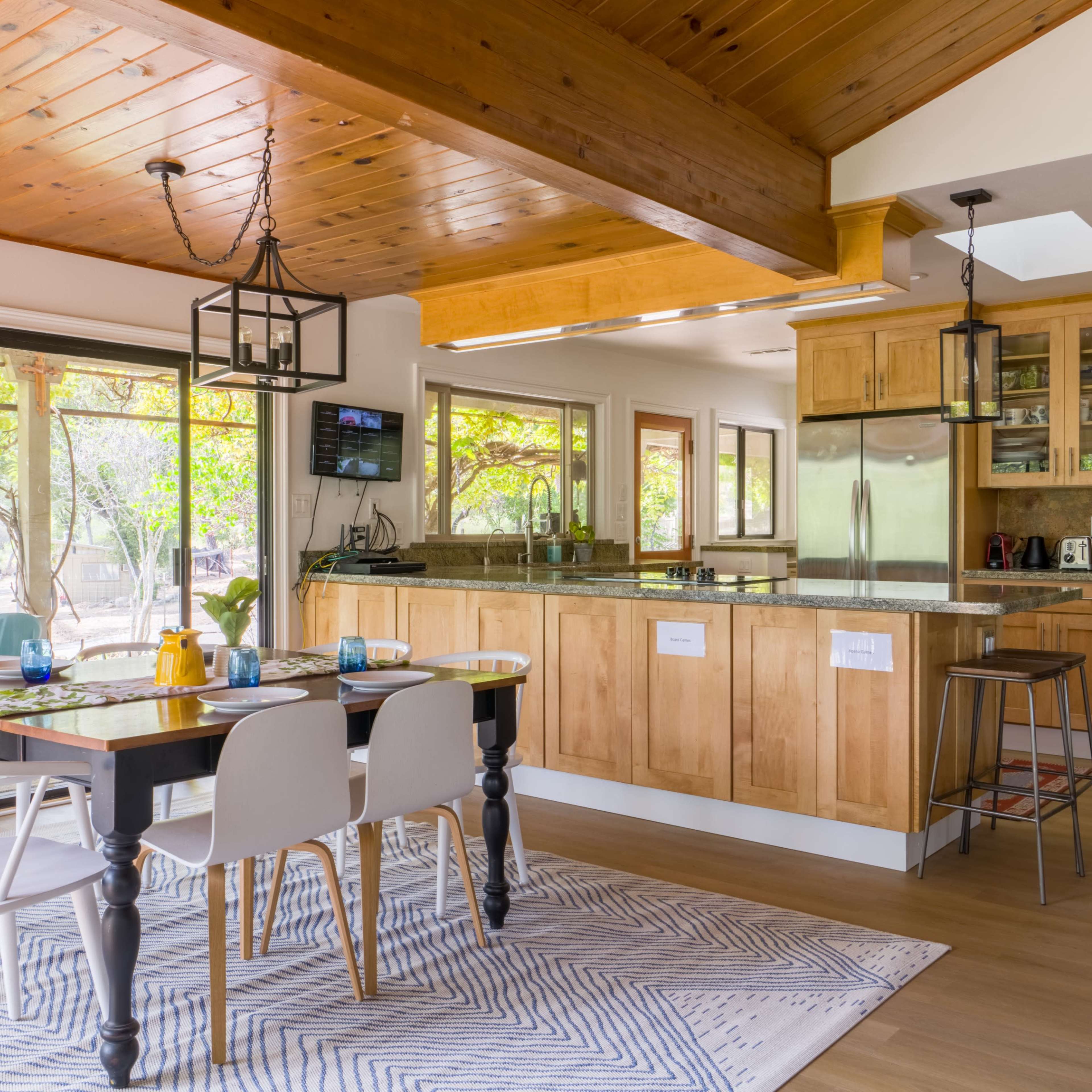 A modern kitchen and dining area featuring wood cabinetry, a central island, a dining table with white and black chairs, and large windows letting in natural light.