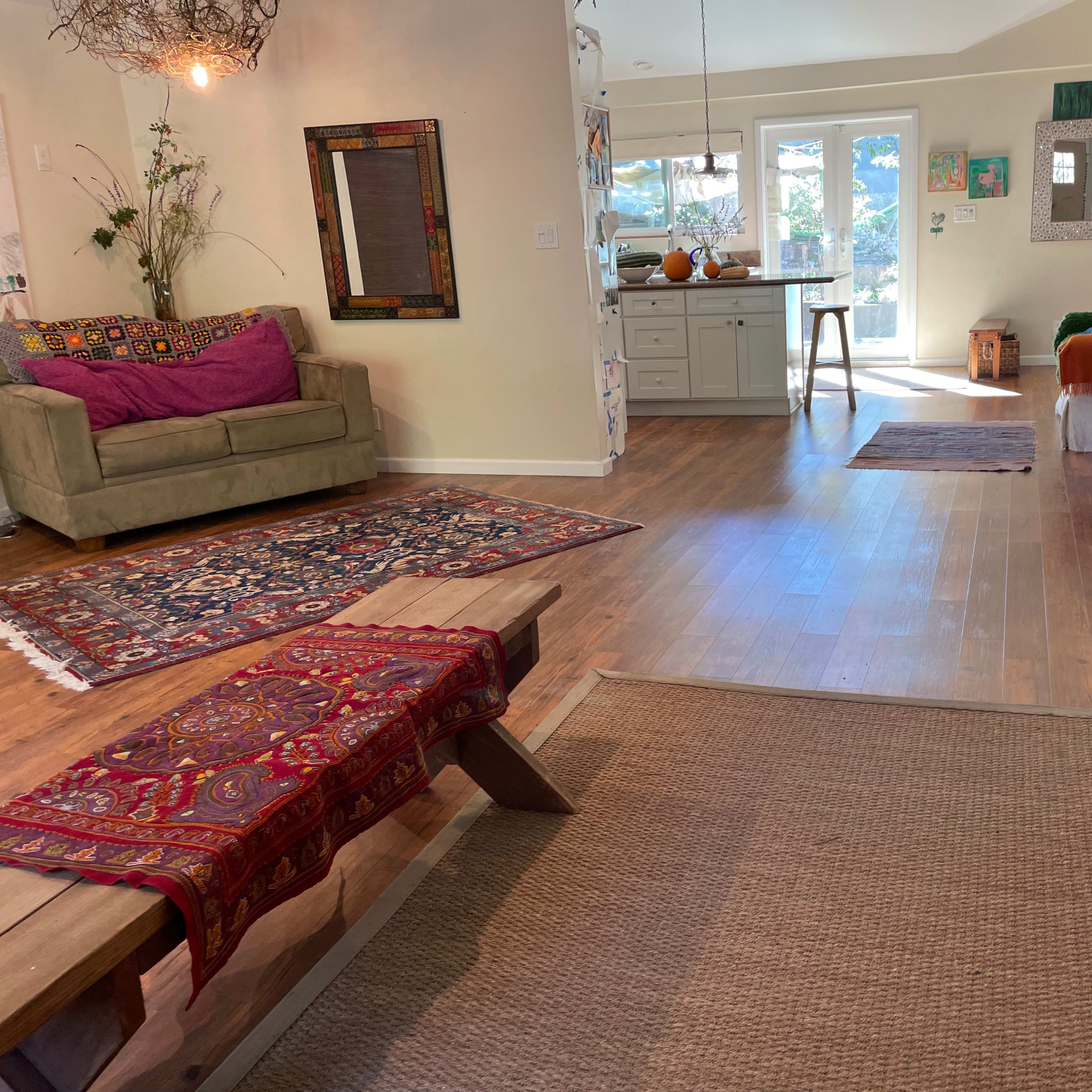A spacious living area featuring a green sofa, decorative rugs, and an open kitchen area with sunlight streaming through large windows.