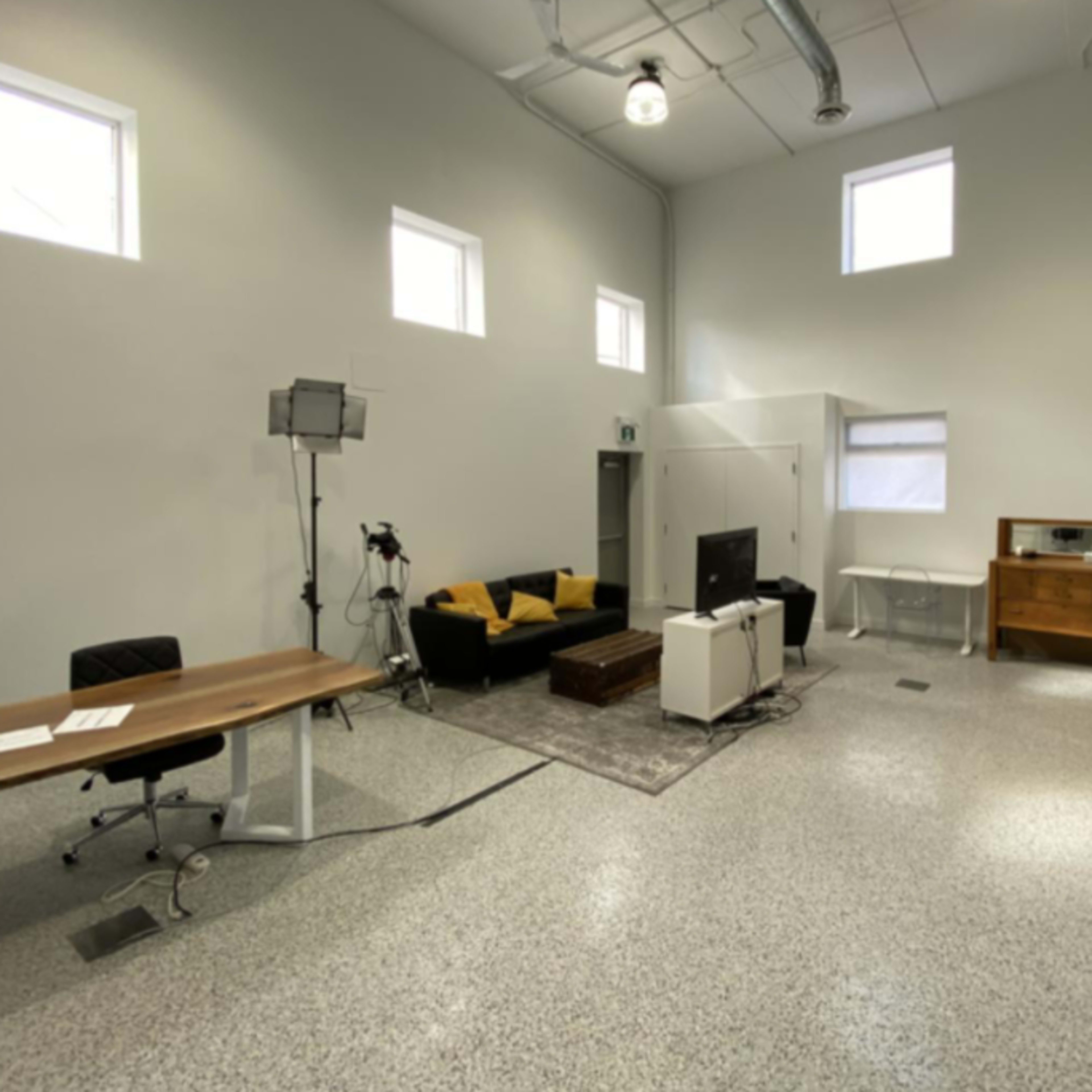 A spacious, modern room with a table, chairs, a couch, and a television, illuminated by natural light from several windows.