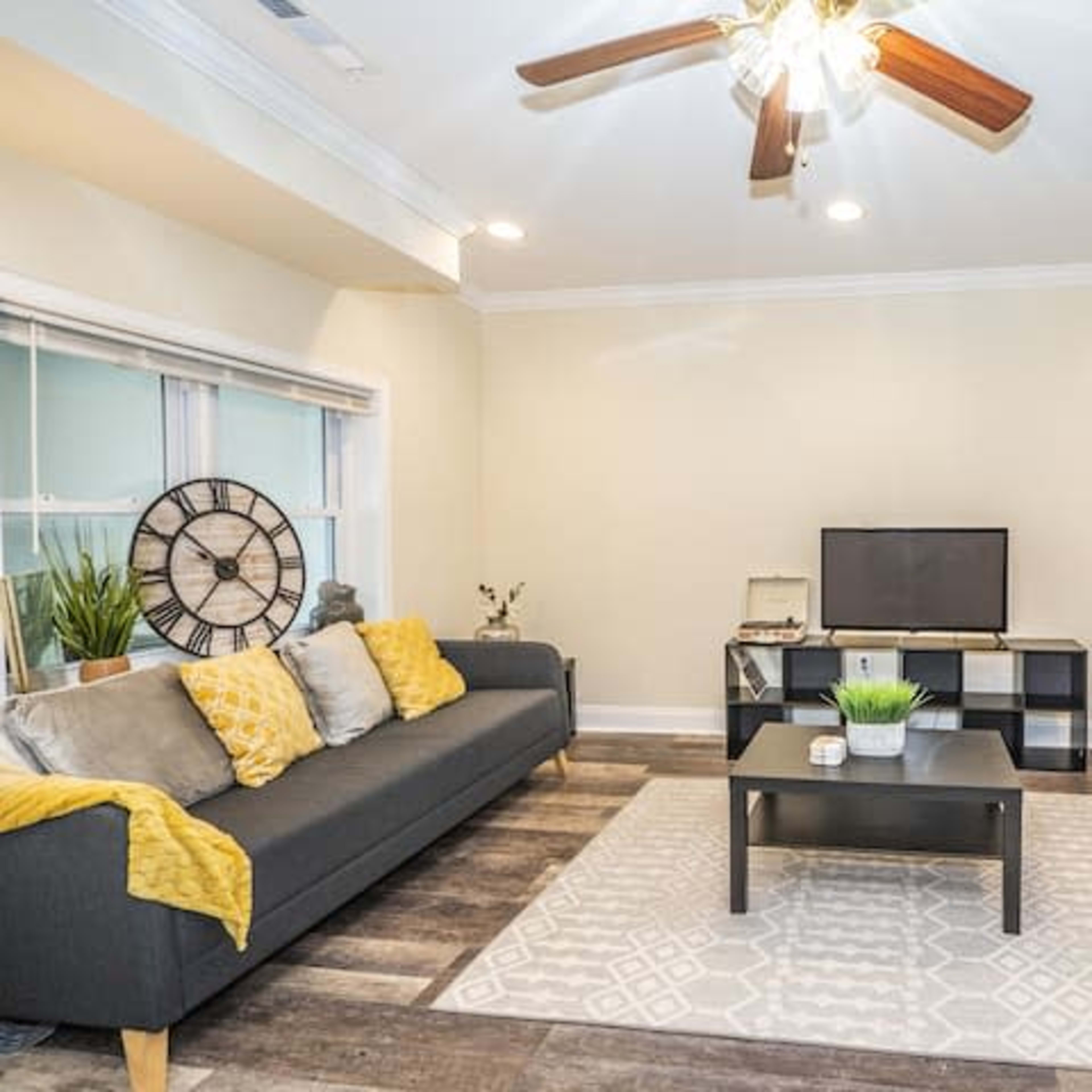 A living room features a dark sofa with yellow throw pillows, a coffee table, and a television on a stand against a light-colored wall.