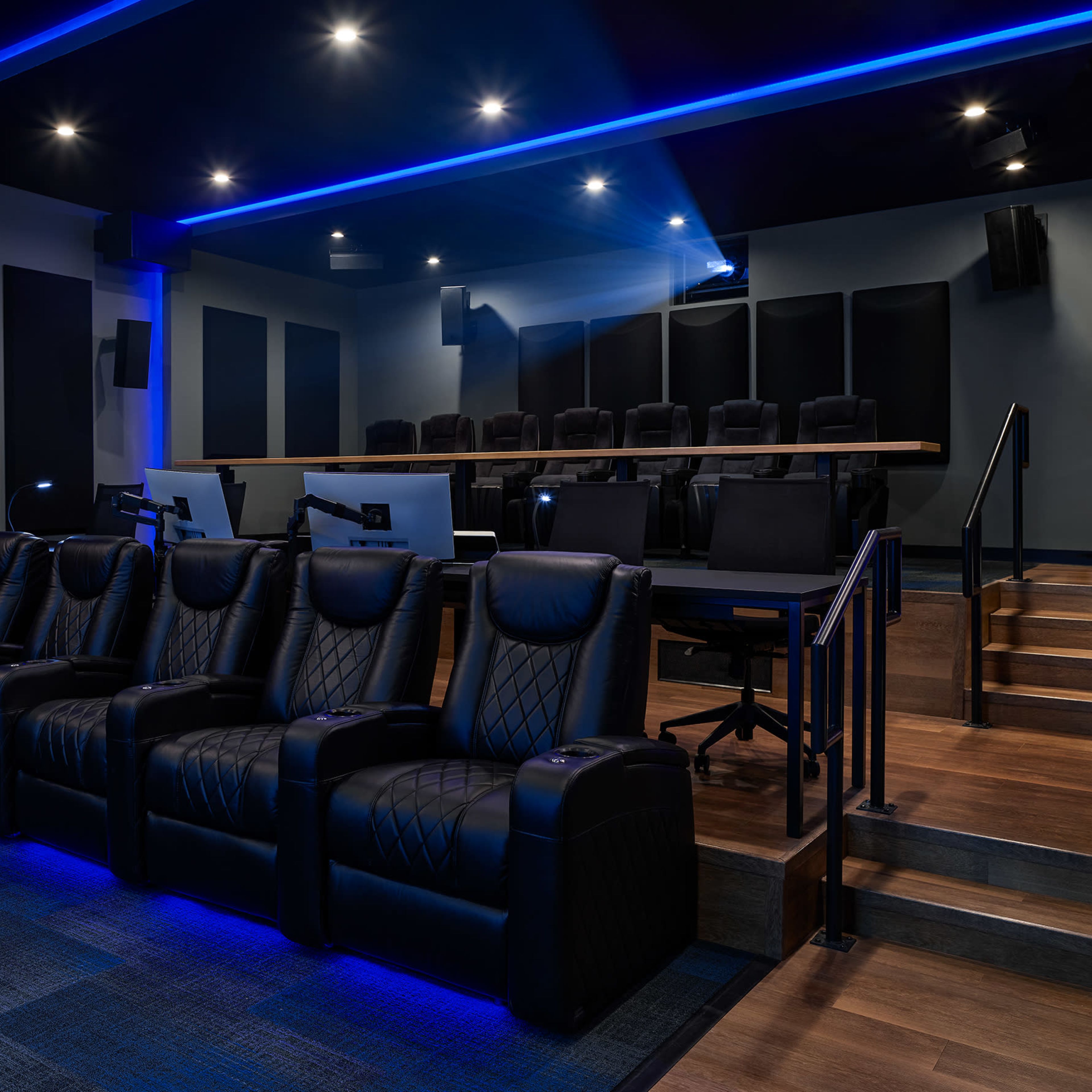 A modern home theater with black recliner seats arranged in front of a tiered seating area and ambient blue lighting.