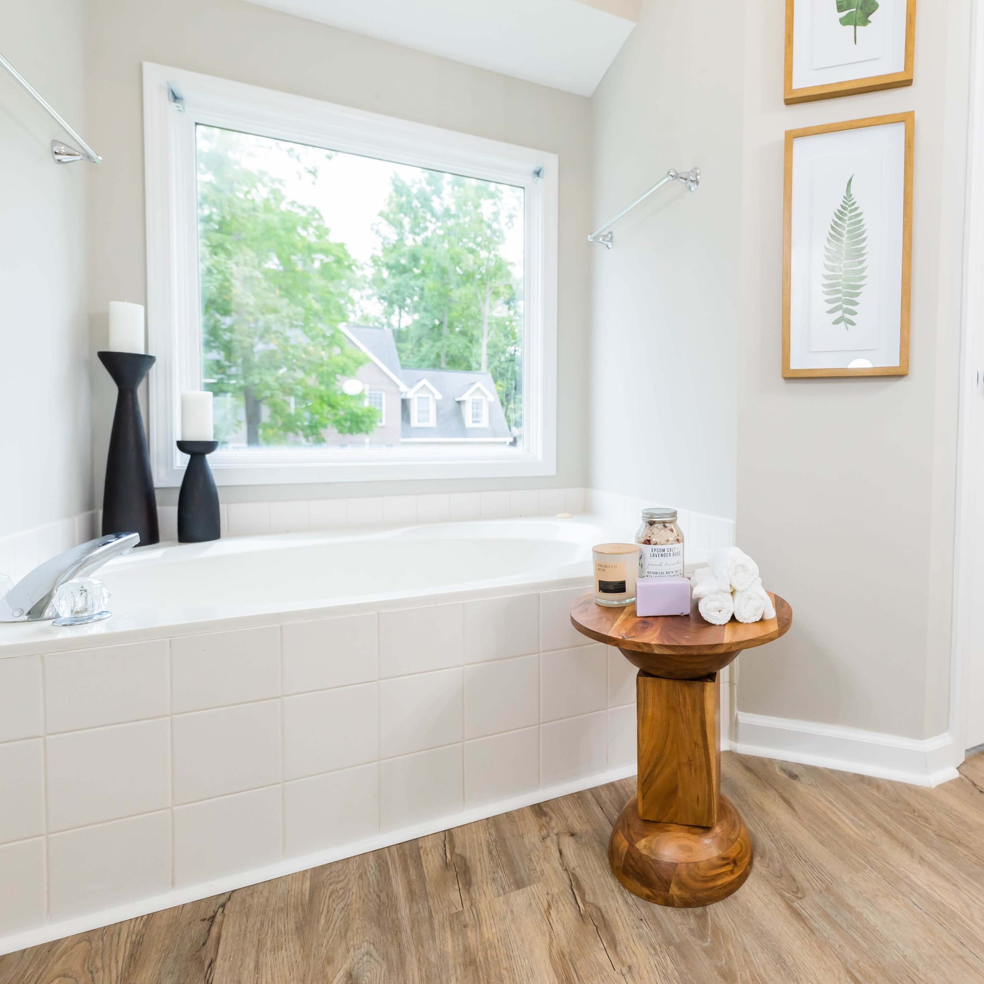 A modern bathroom features a bathtub beside a wooden side table holding towels and toiletries, with a window providing natural light.