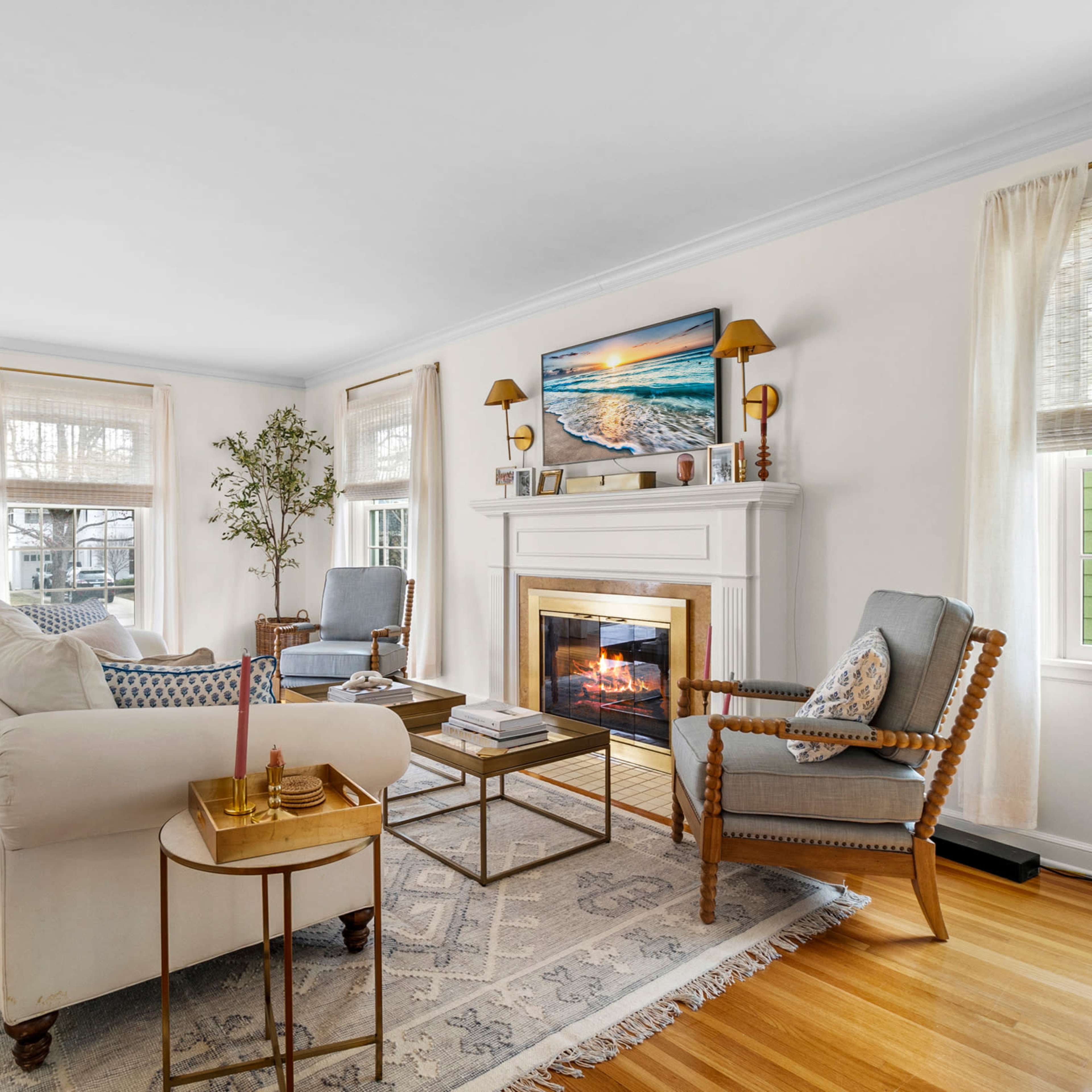 The bright living room features a cozy fireplace, two armchairs, a sofa, and windows allowing natural light to illuminate the wood flooring.