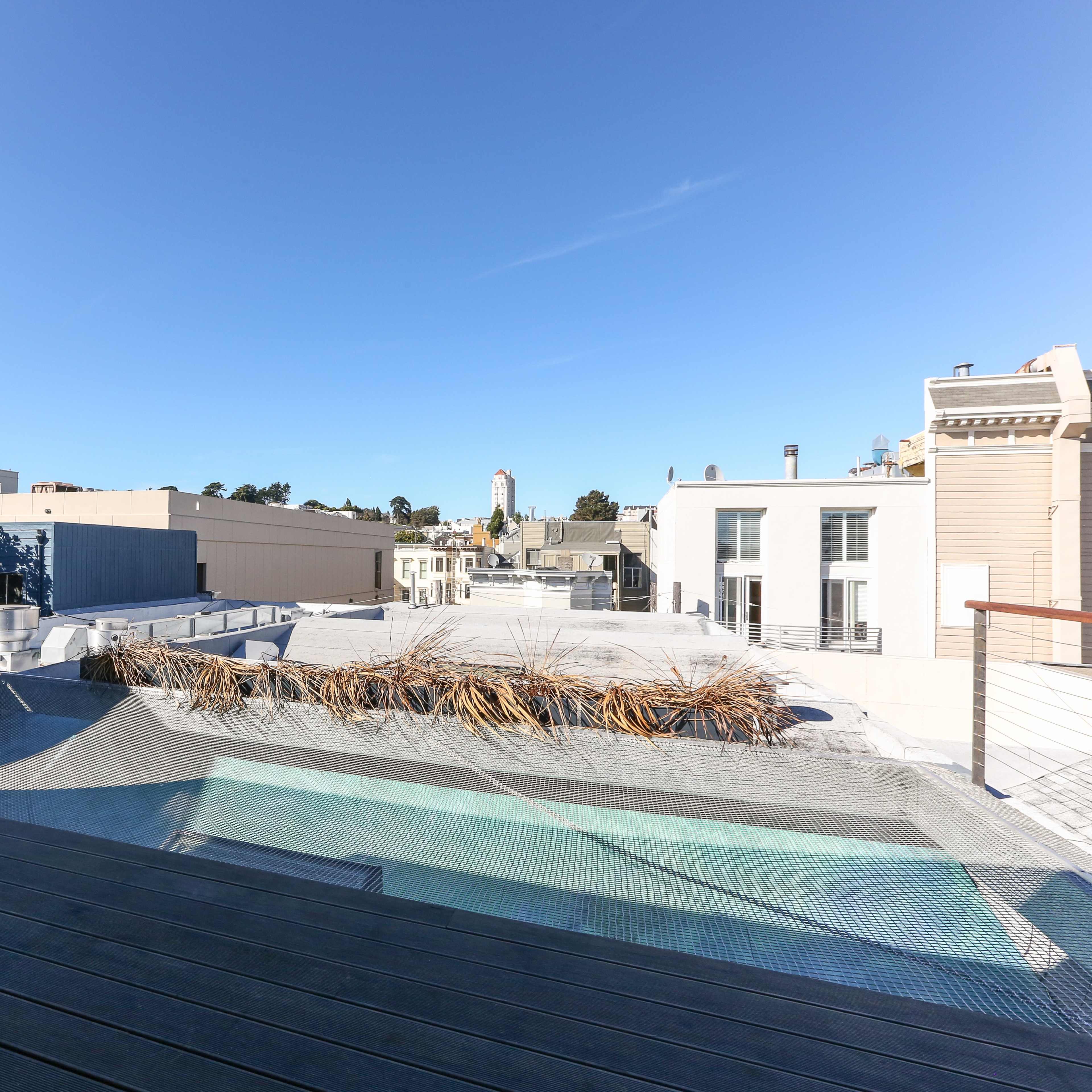 A rooftop deck with a glass railing overlooks city buildings and clear blue skies.