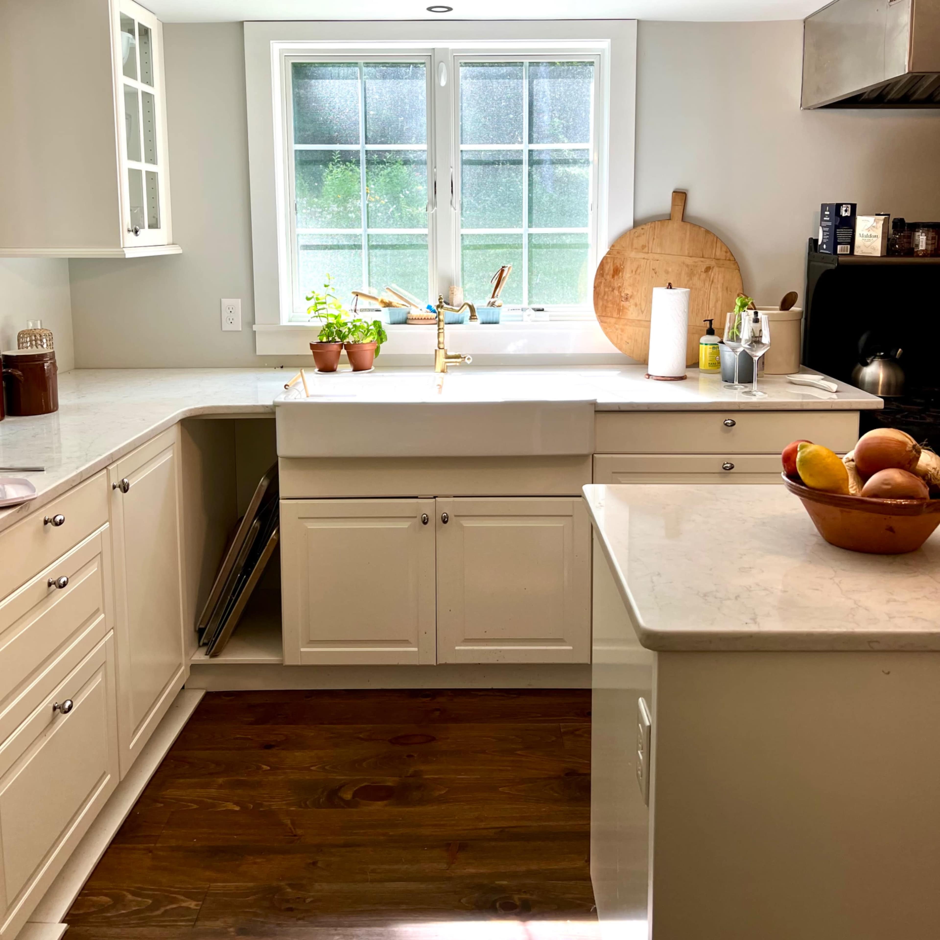 A bright kitchen features white cabinetry, a large window above the sink, and potted plants on the countertop.