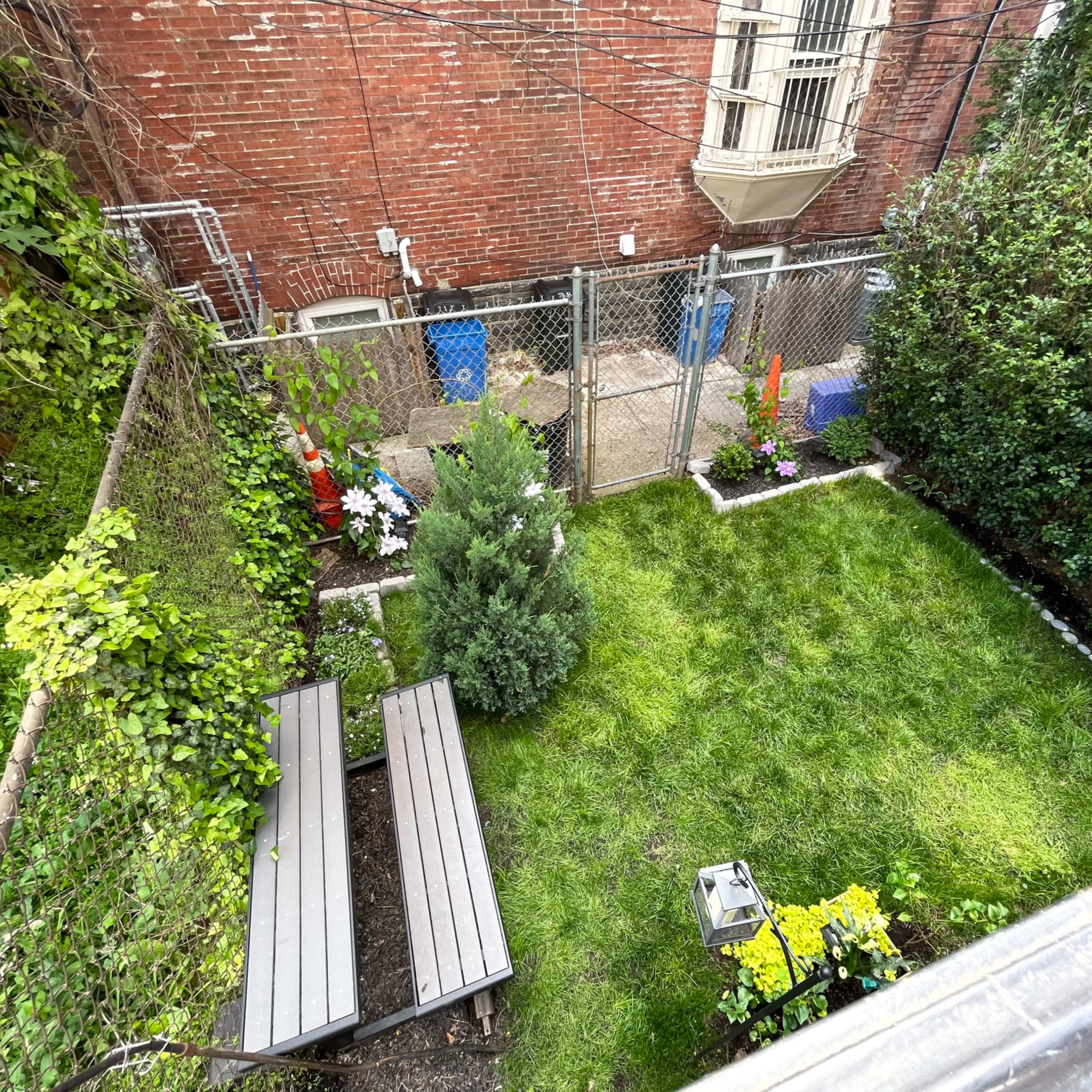 A fenced backyard with a manicured lawn, contains two wooden benches, and is bordered by shrubs and flowers.