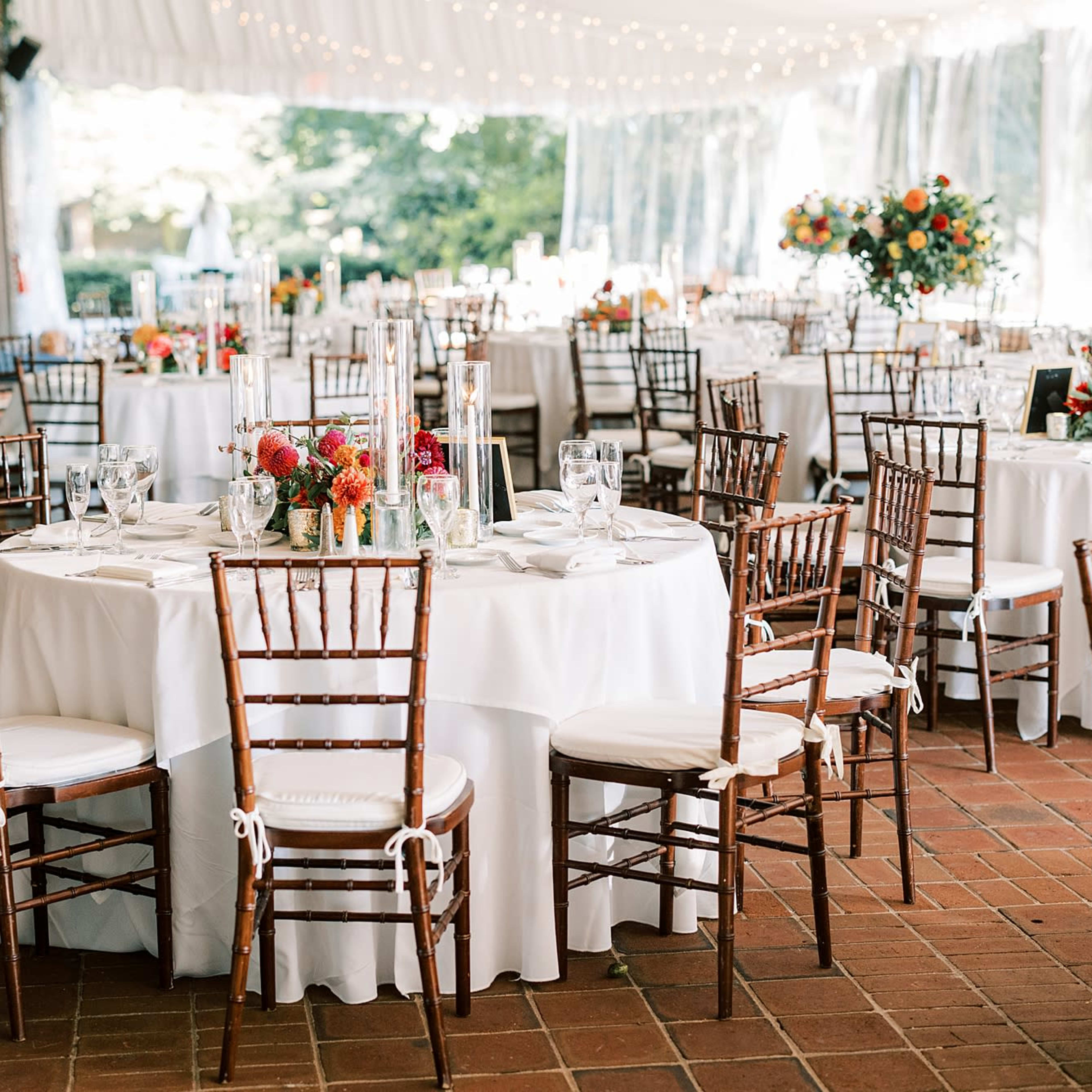 A banquet hall is set up with round tables covered in white tablecloths, surrounded by wooden chairs, and adorned with floral centerpieces.