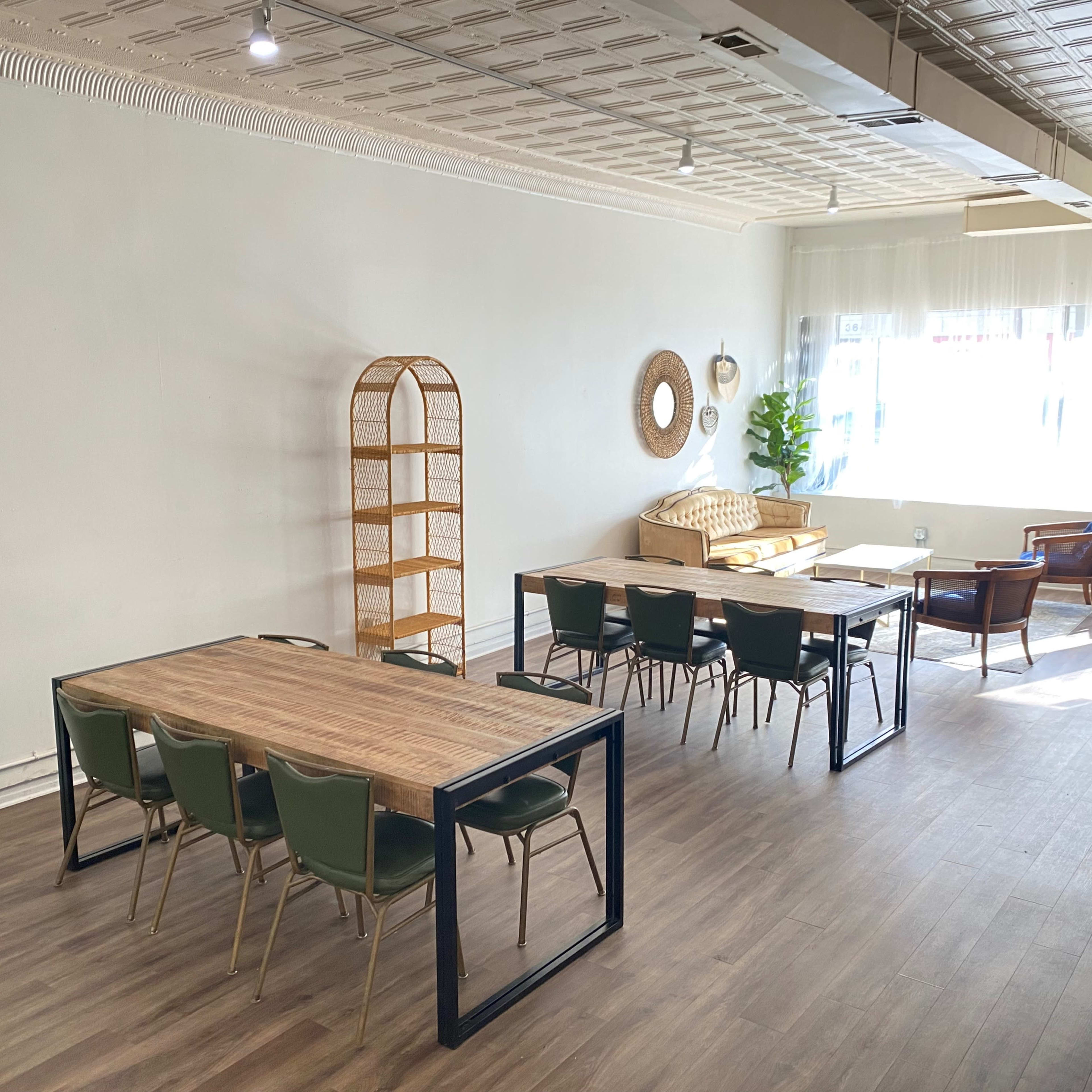 A spacious room with wooden flooring, featuring multiple tables and chairs, a wicker shelf, a sofa, and large windows allowing natural light.