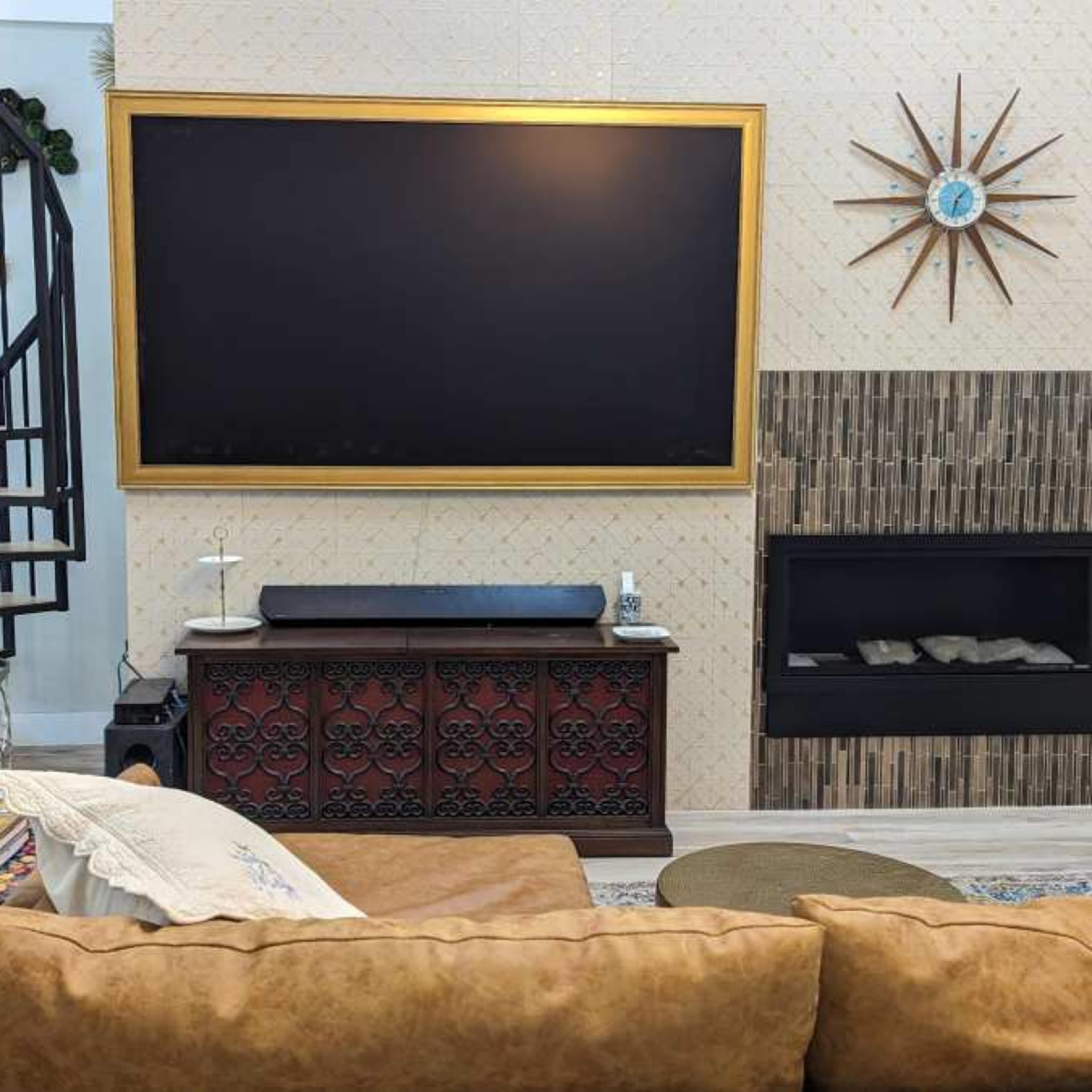A living room with a large wall-mounted TV framed in gold, a decorative wall clock, a spiral staircase, and a modern fireplace below a TV unit.