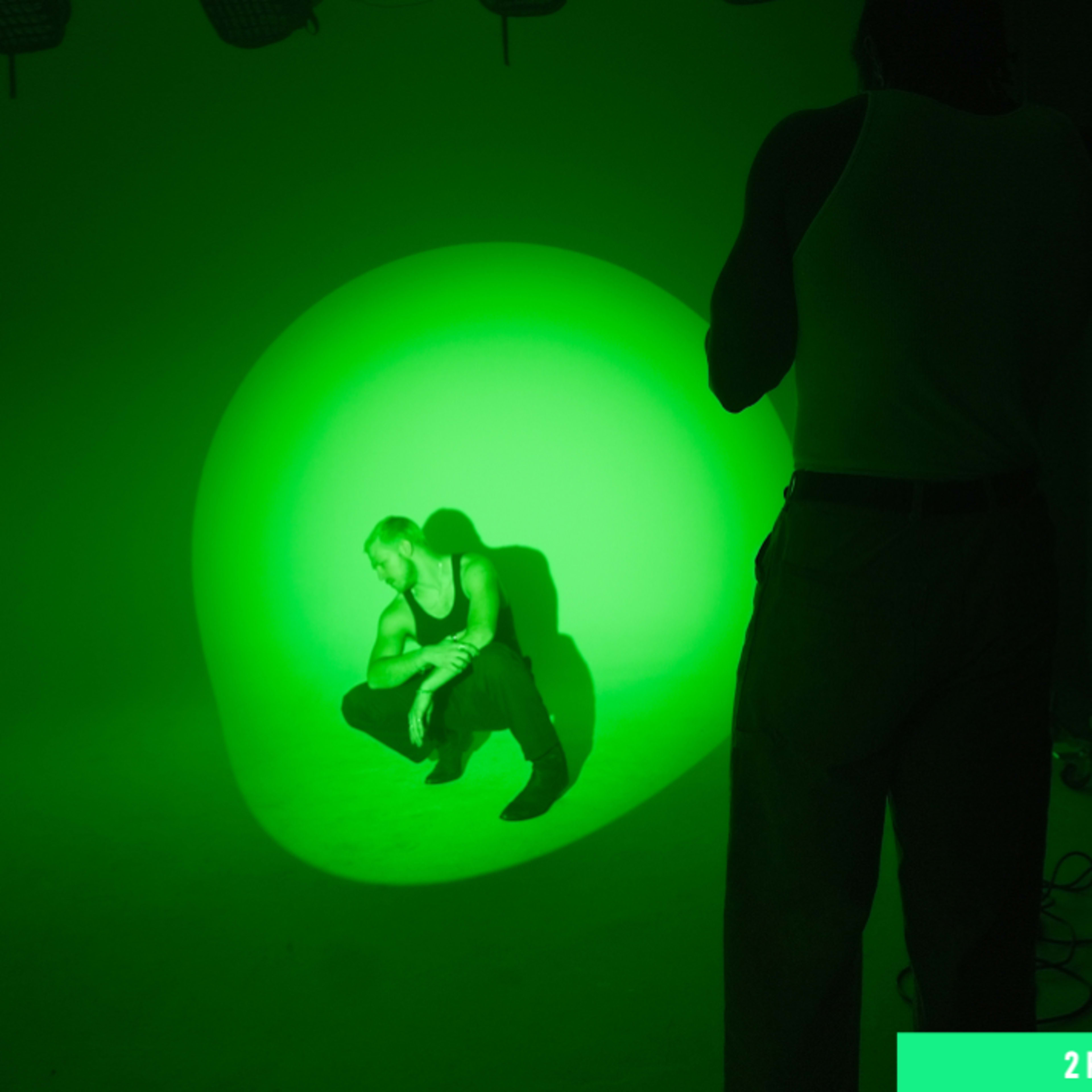 A person crouches in a green-lit studio while another figures are positioned behind a camera.