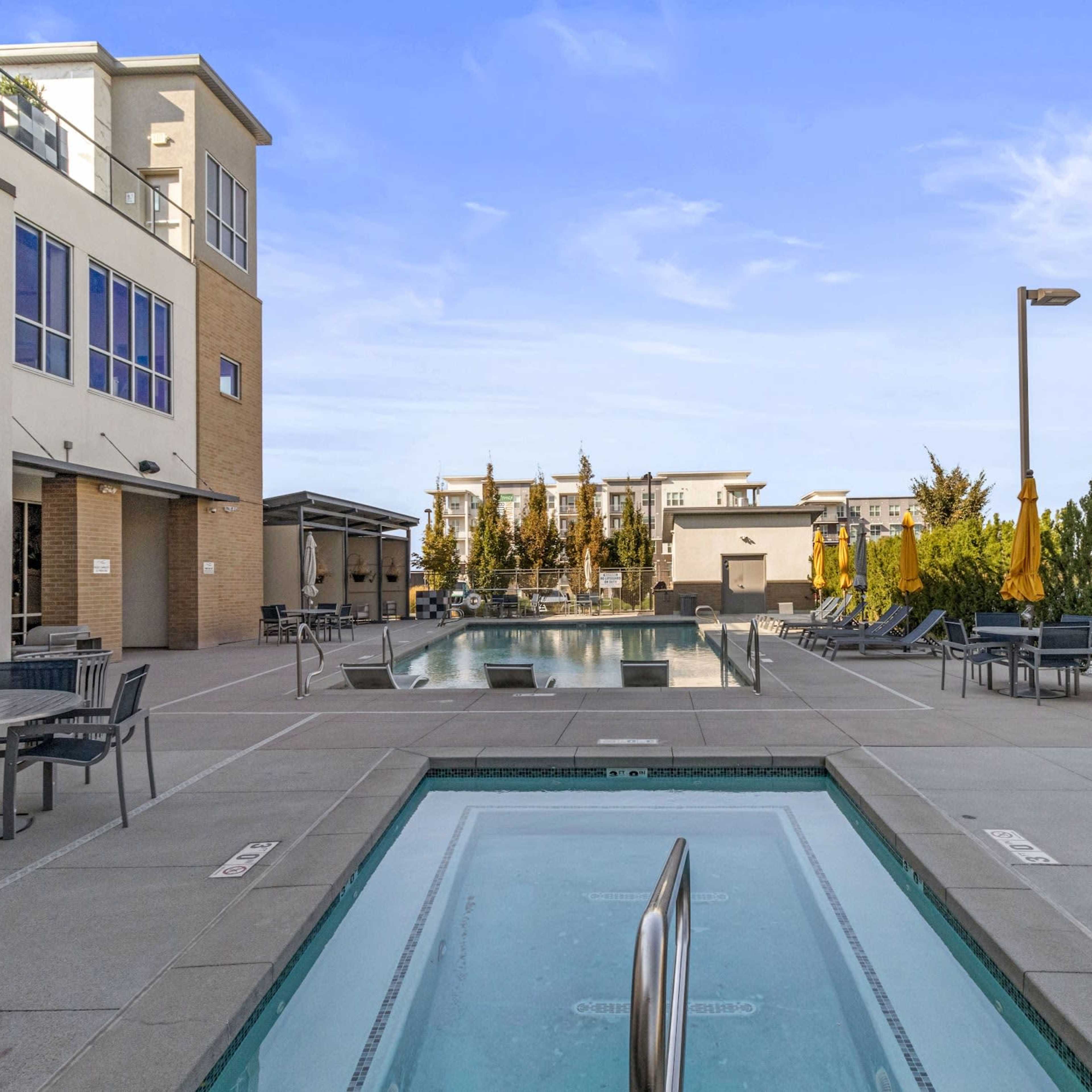 A modern outdoor swimming pool area with lounge chairs, umbrellas, and surrounding greenery.