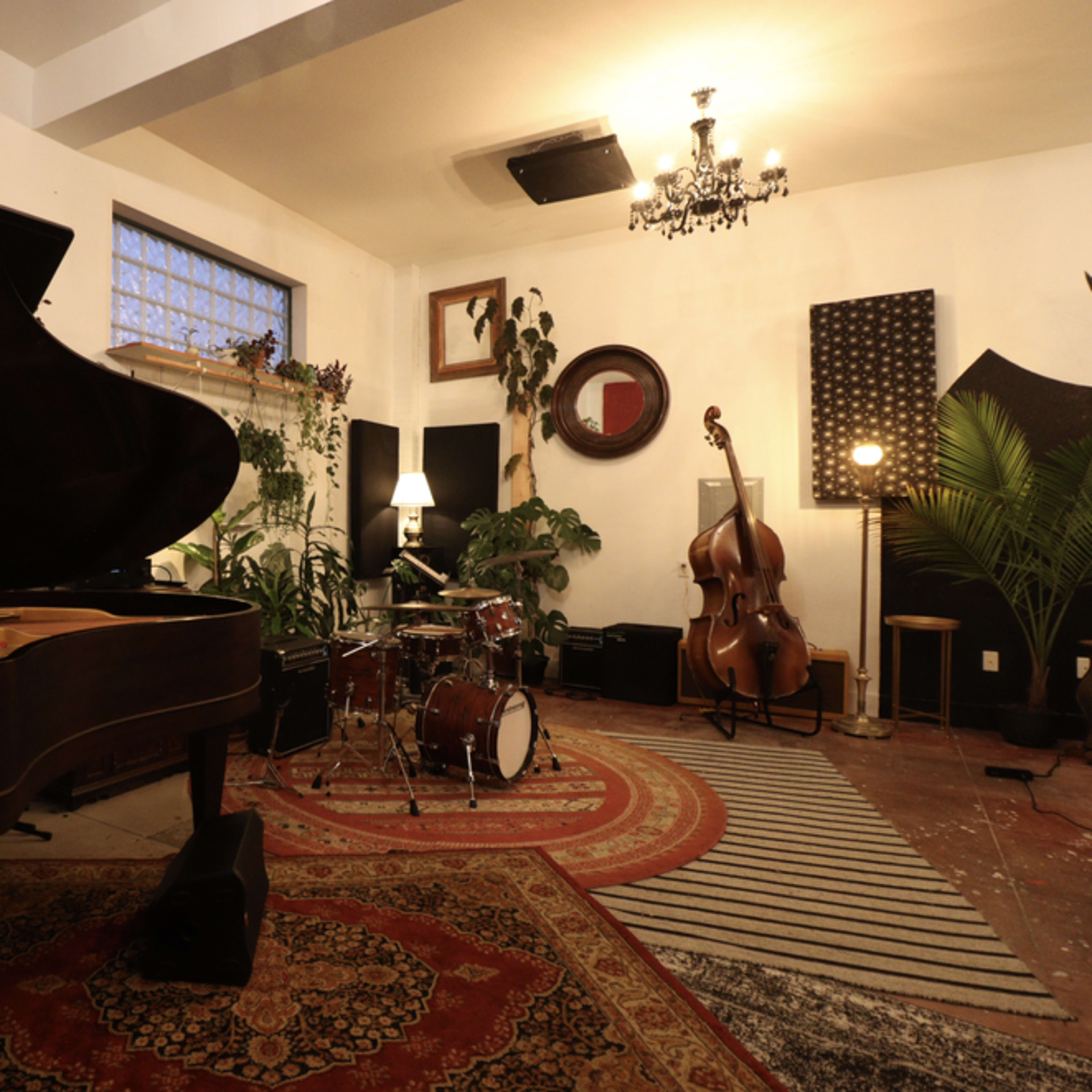 The room features a grand piano, a drum set, a double bass, and lush plants, all arranged around a central area on a patterned rug.