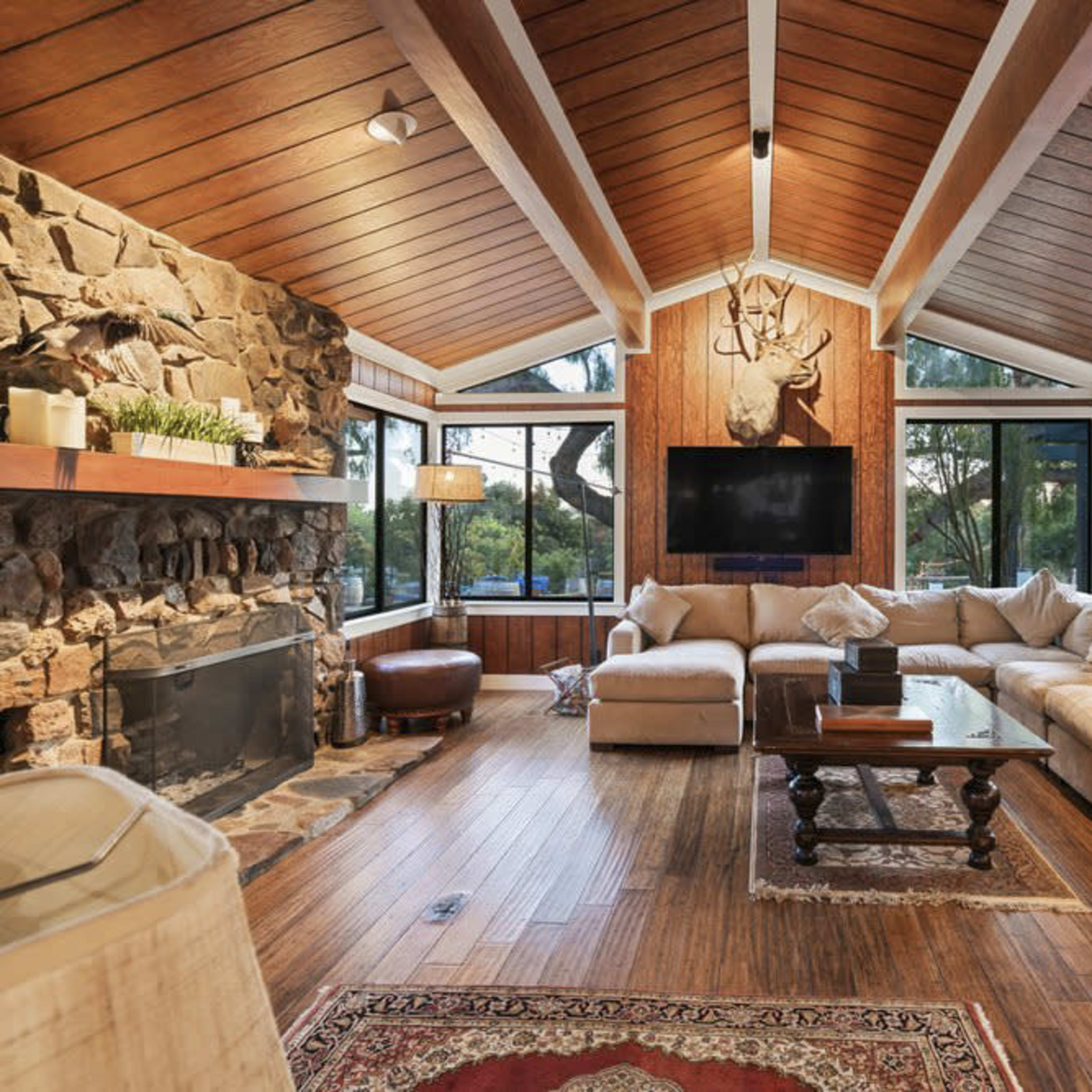 A cozy living room with wood-paneled walls, a stone fireplace, and a large sectional sofa arranged around a wooden coffee table.