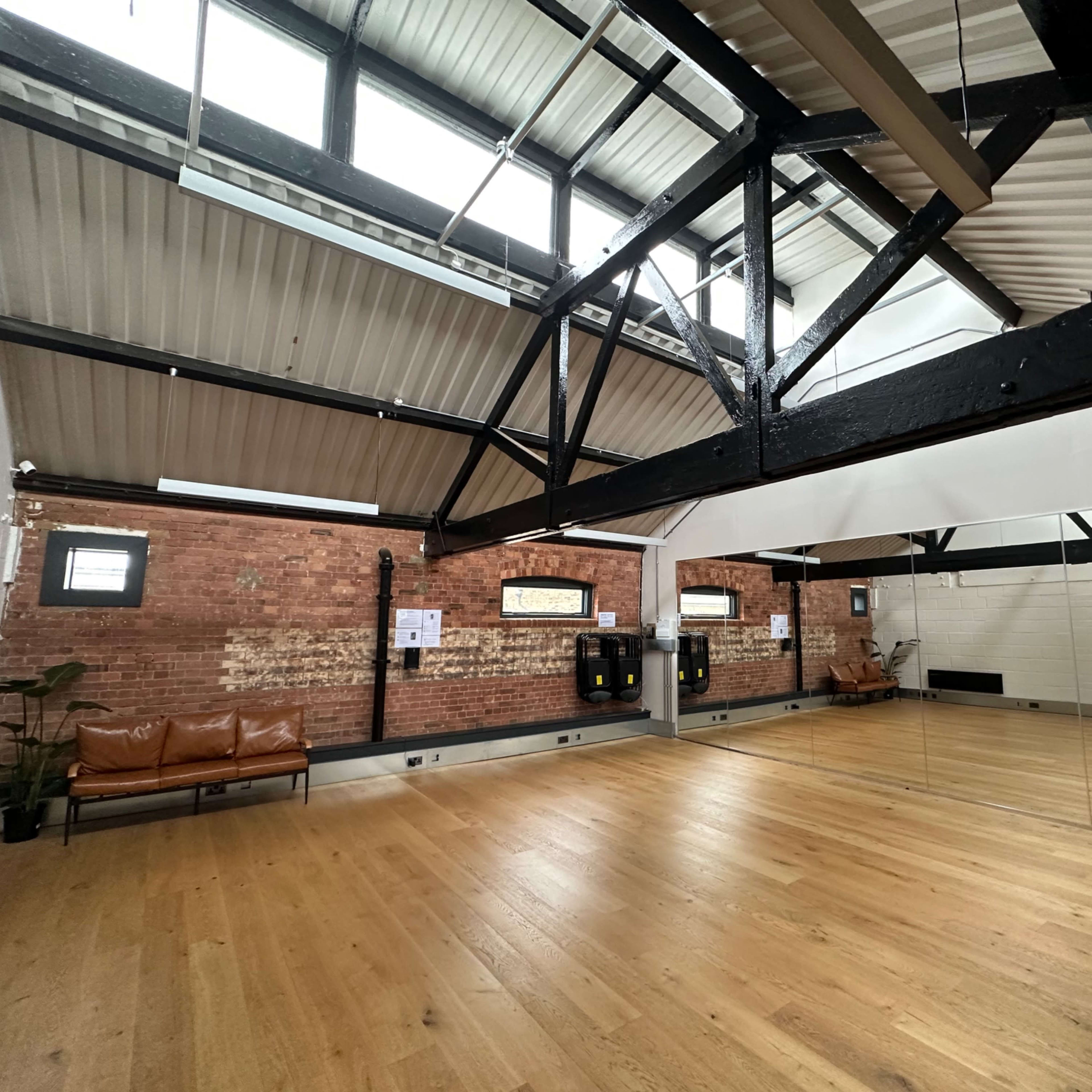 An interior of a spacious studio with wooden flooring, exposed brick walls, and high ceilings supported by black beams.