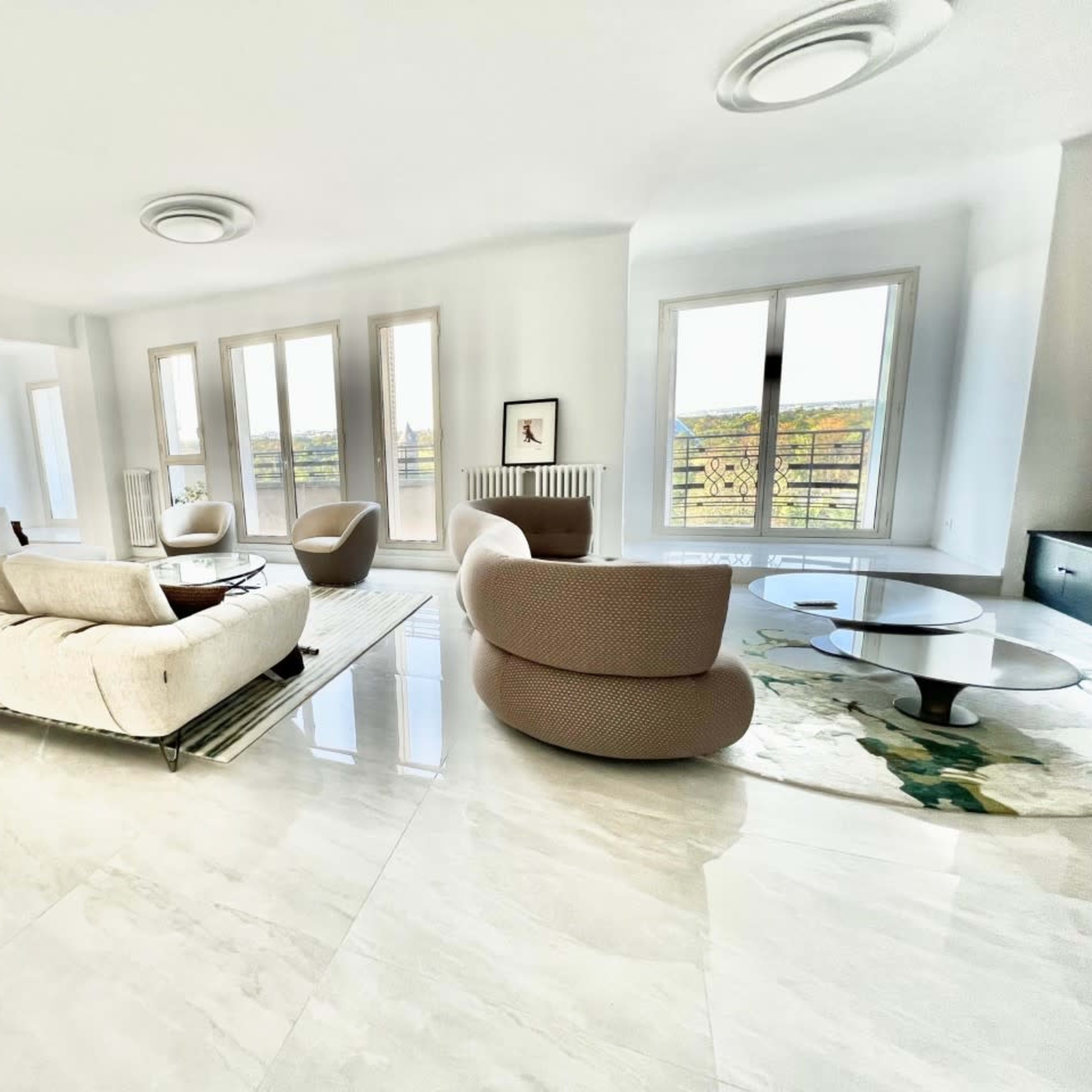 A spacious living room featuring modern furniture, large windows, and a marble floor.