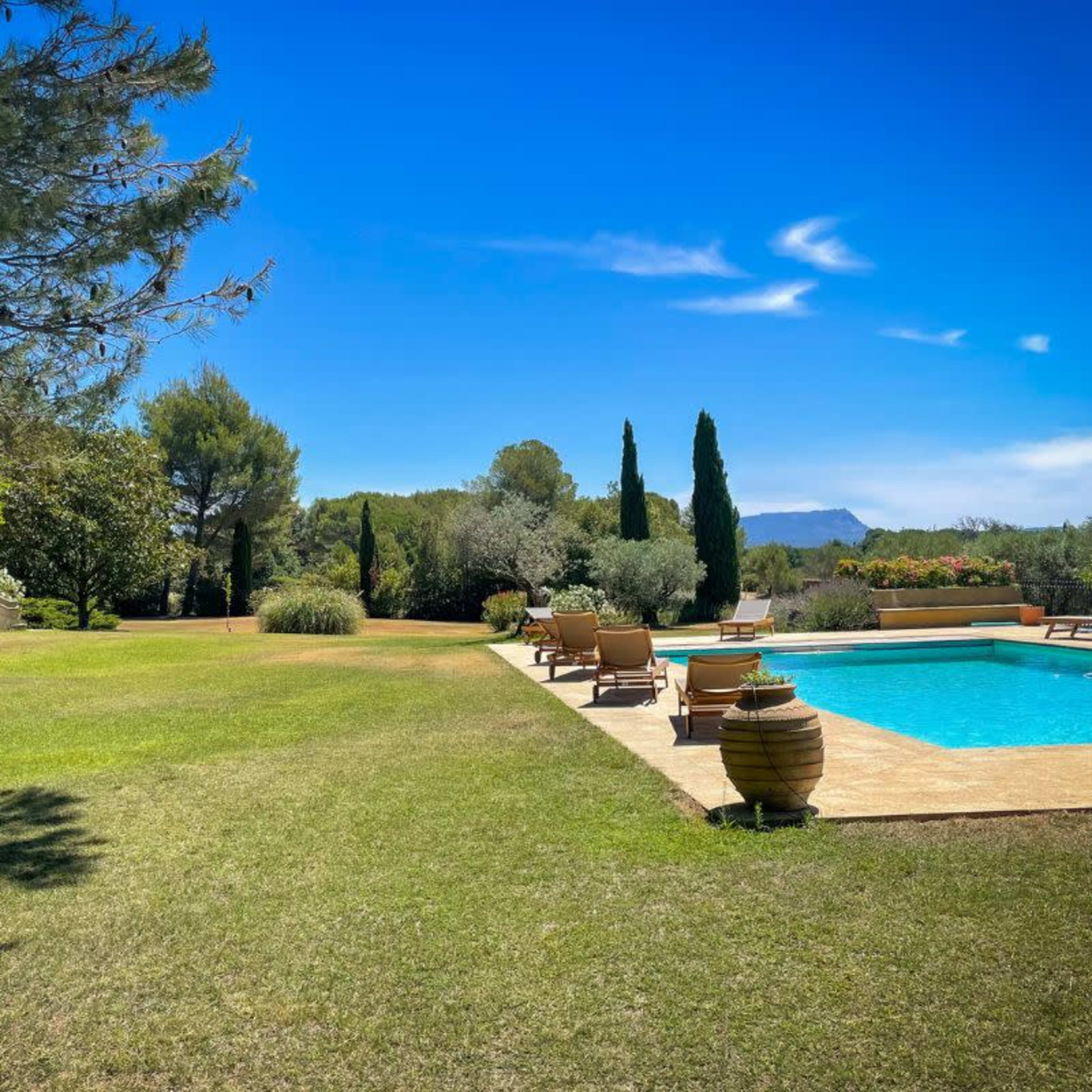 A landscaped backyard featuring a swimming pool surrounded by lounge chairs, with trees and a distant mountain in the background under a clear blue sky.