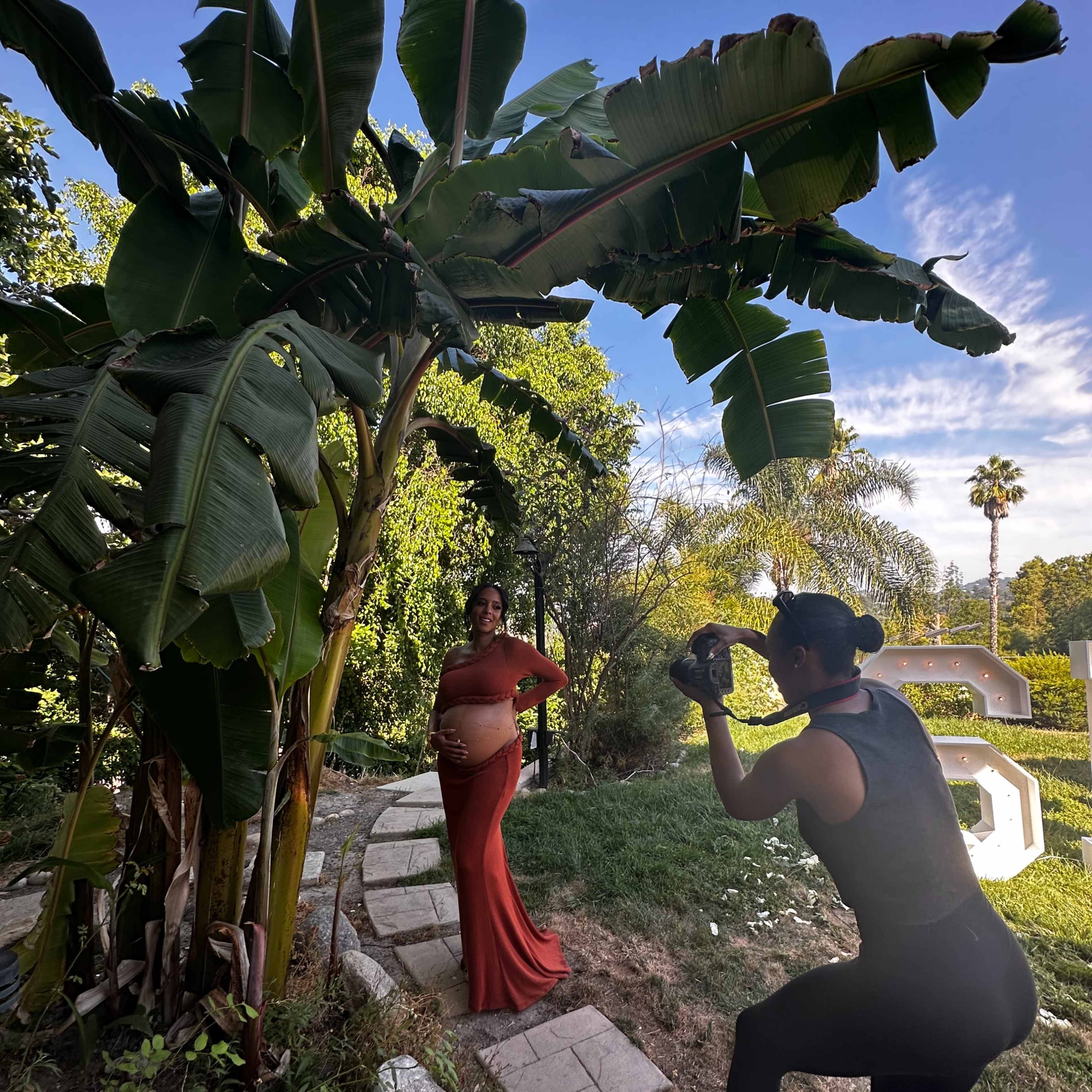 A pregnant woman in a red dress poses for a photo in a garden while a photographer captures the moment.