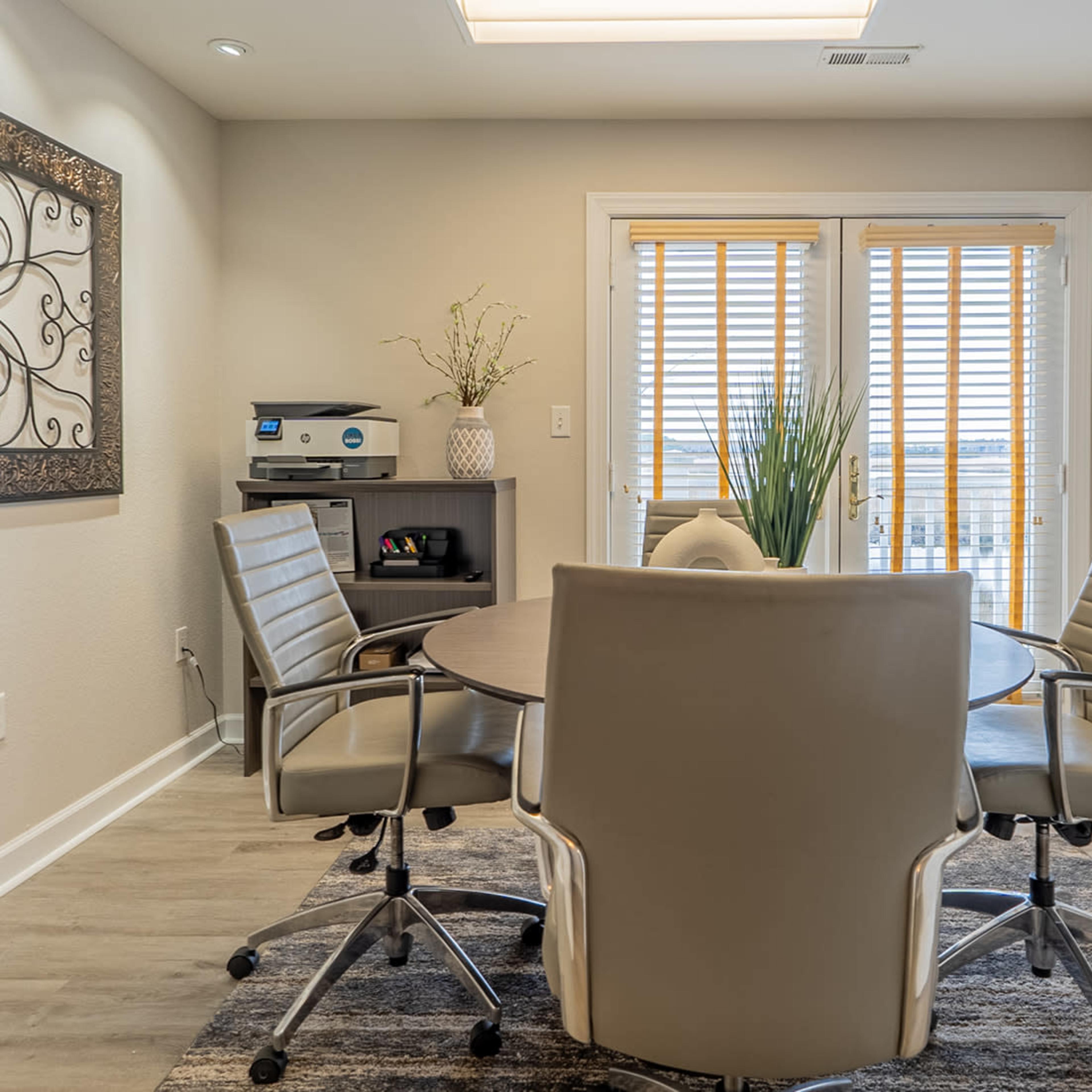A modern meeting room features a round table surrounded by four ergonomic chairs, with a decorative wall, a storage unit, and large windows with blinds.