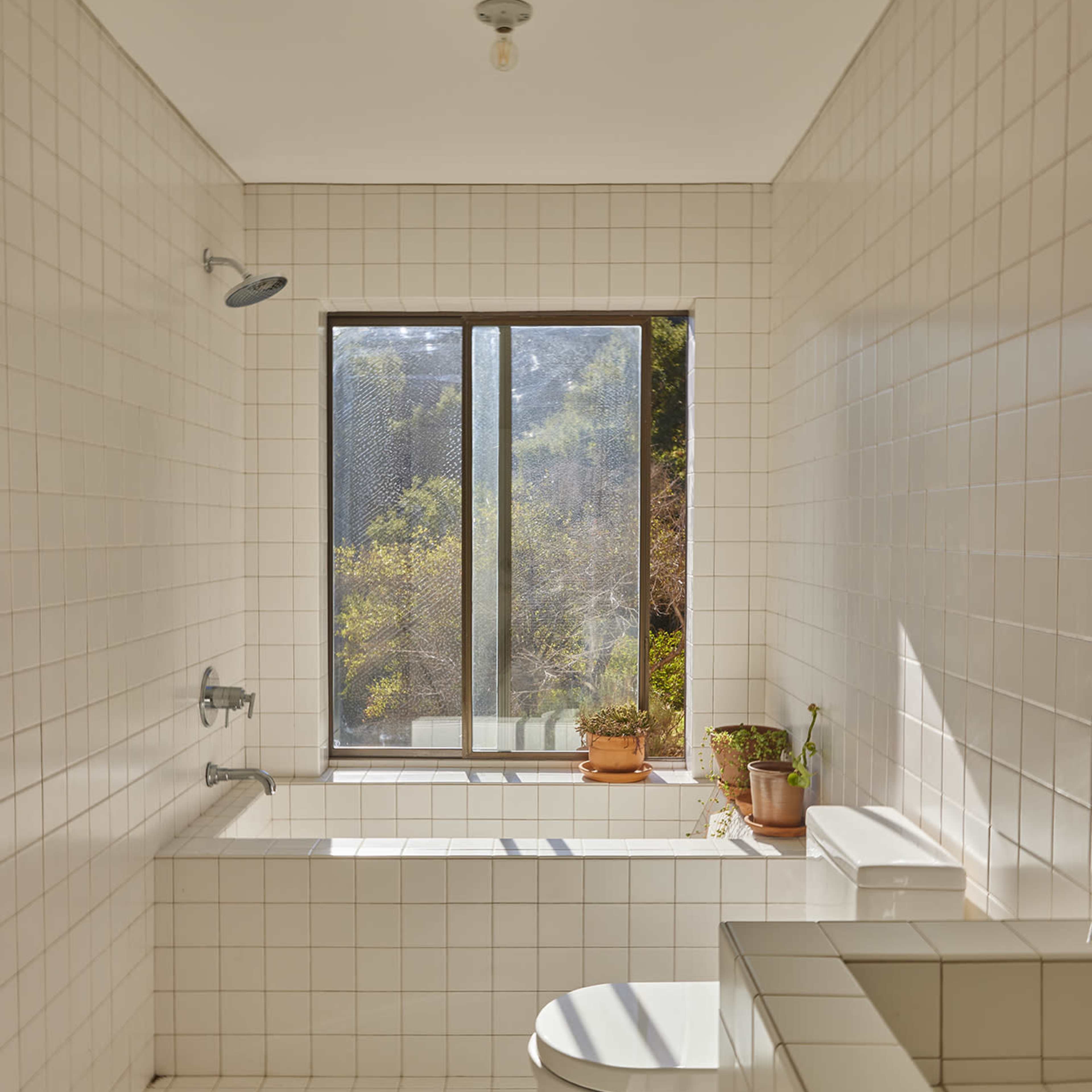 A bright, tiled bathroom features a bathtub next to a large window with plants on the sill.