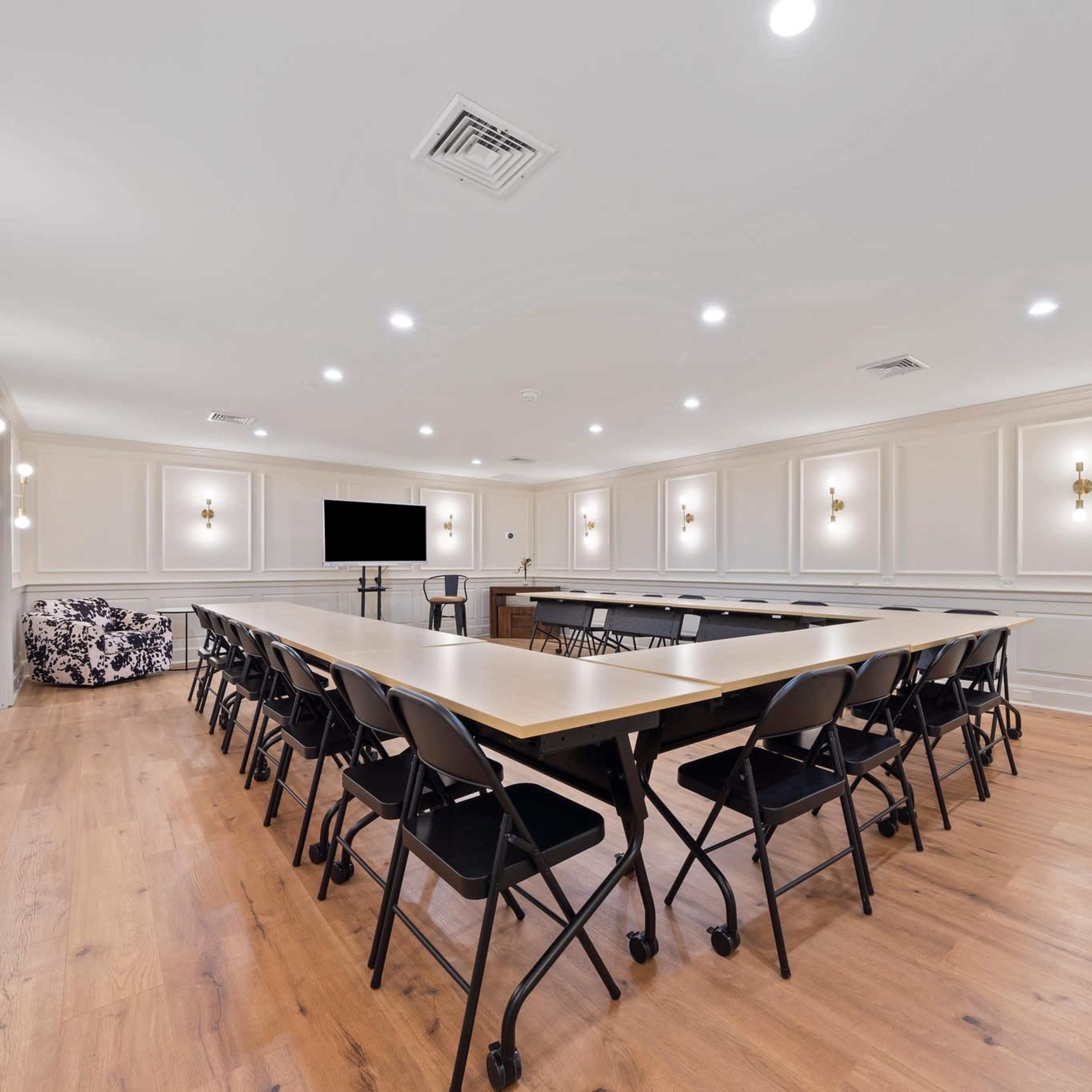 A spacious meeting room with a large central table surrounded by black folding chairs, illuminated by wall sconces and featuring a television screen at one end.