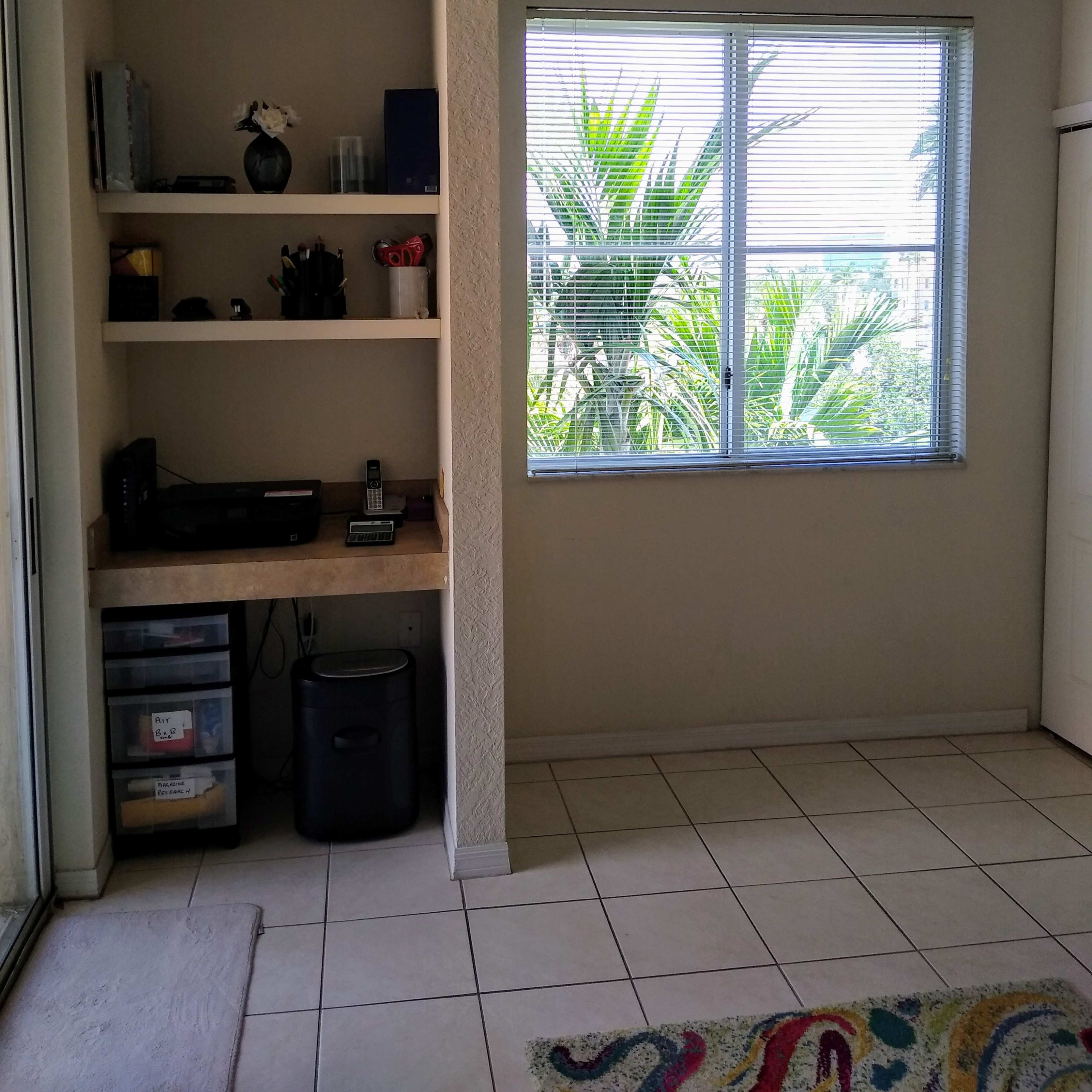 A small, well-lit room features a desk with office supplies and a window overlooking a palm tree landscape.