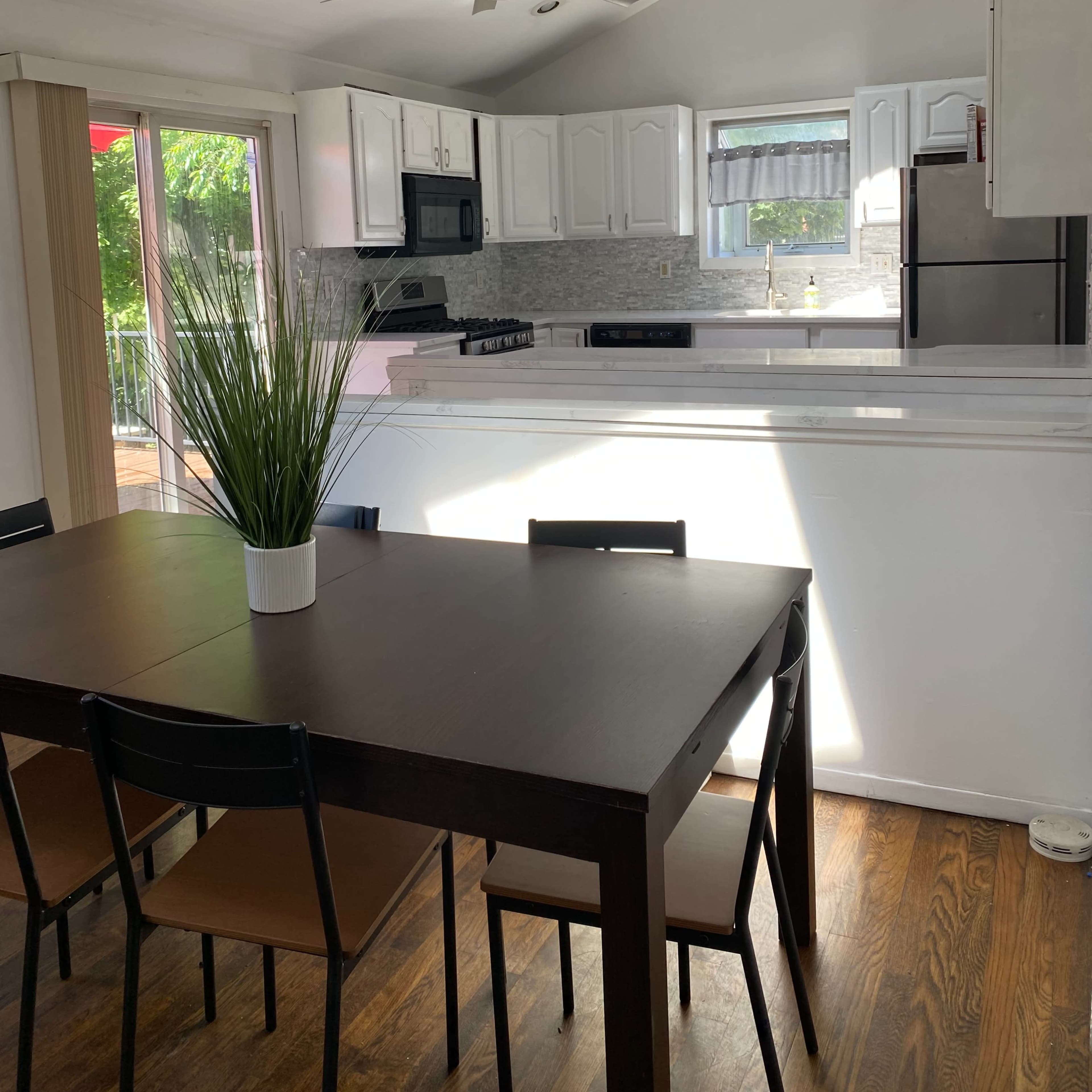 A modern kitchen and dining area with a table, black chairs, and a ceiling fan, featuring white cabinetry and a large window.