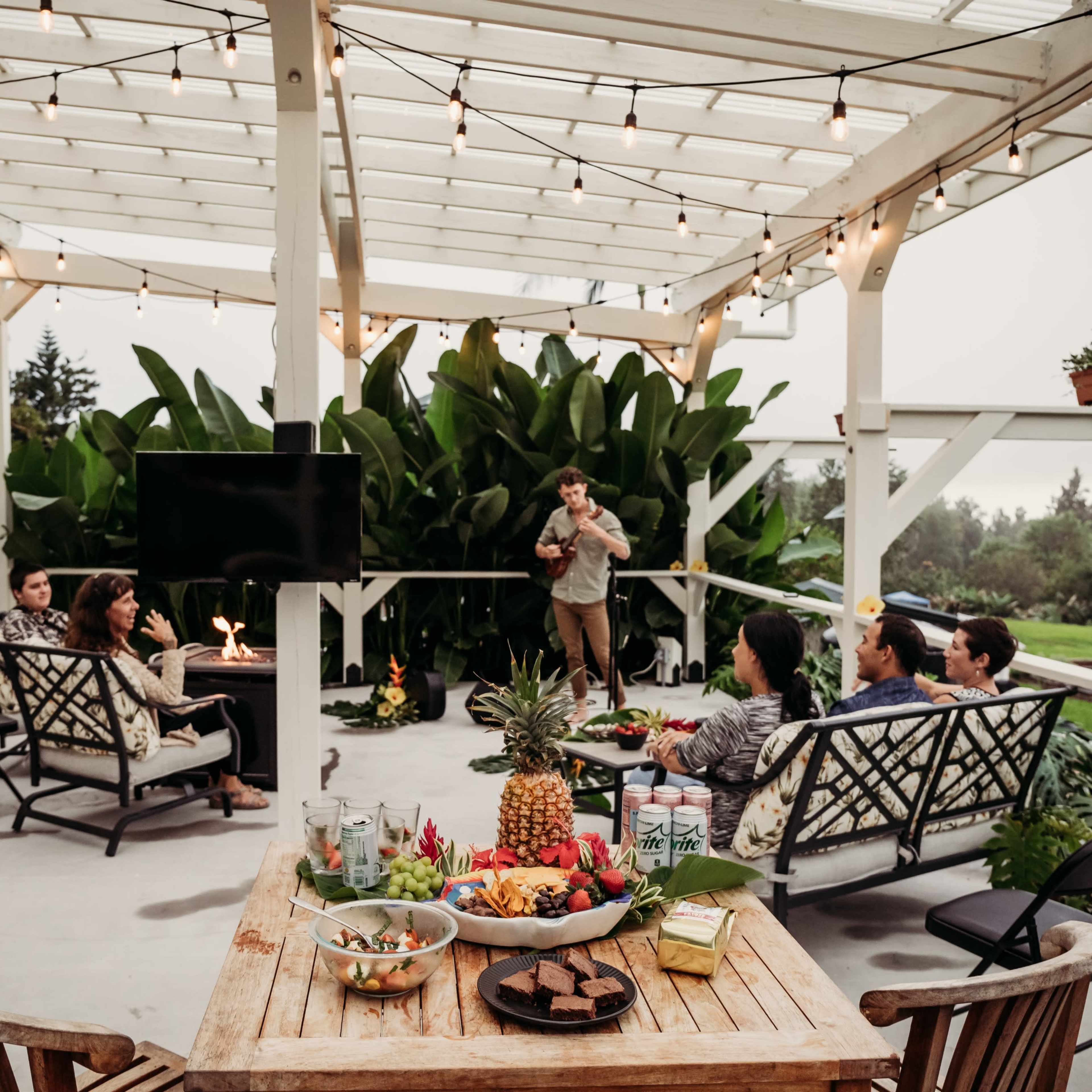 A group of people gathers on a covered patio with string lights, enjoying food and drinks, while someone plays music in front of them.