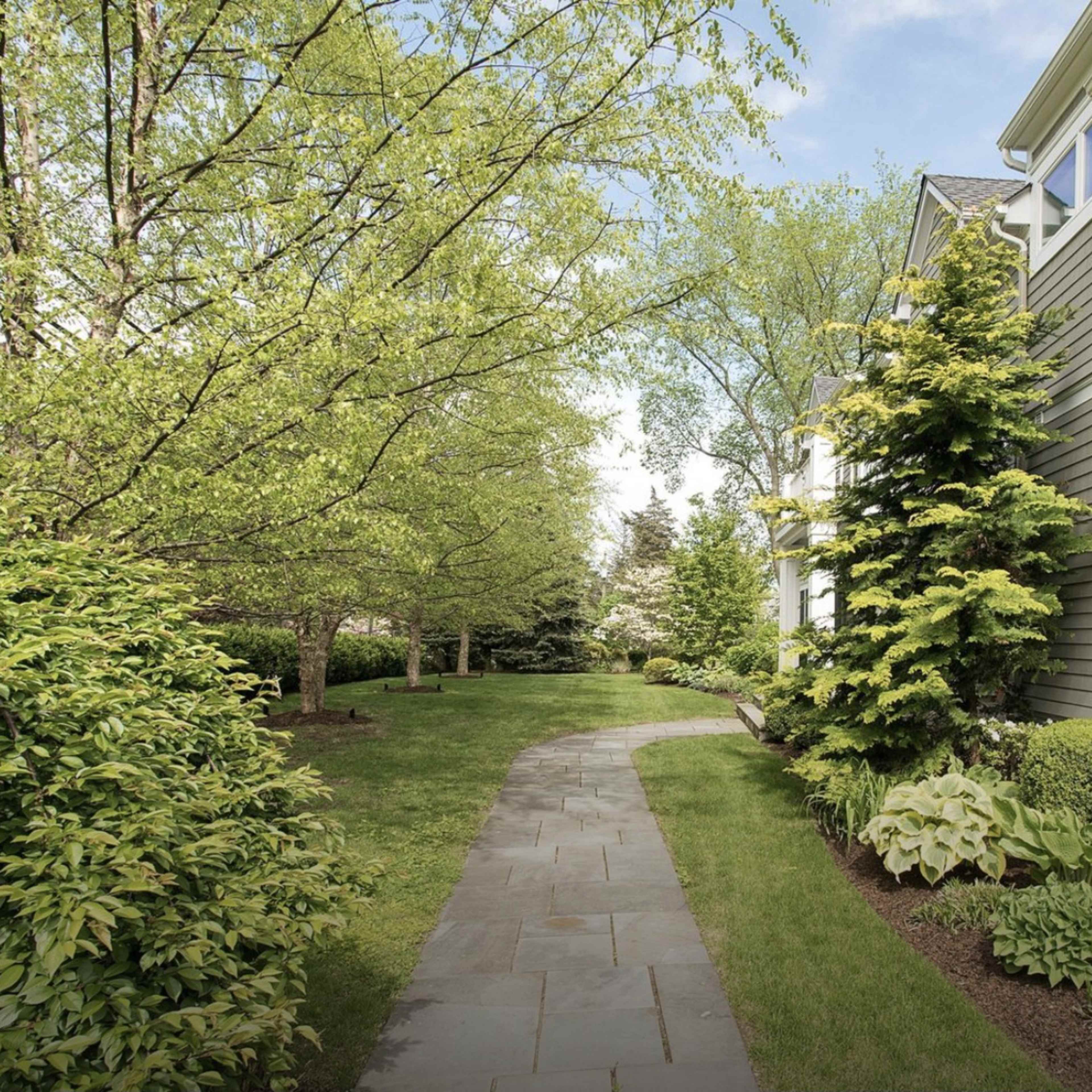 A stone pathway curves alongside a manicured lawn bordered by trees and shrubs beside a gray house.