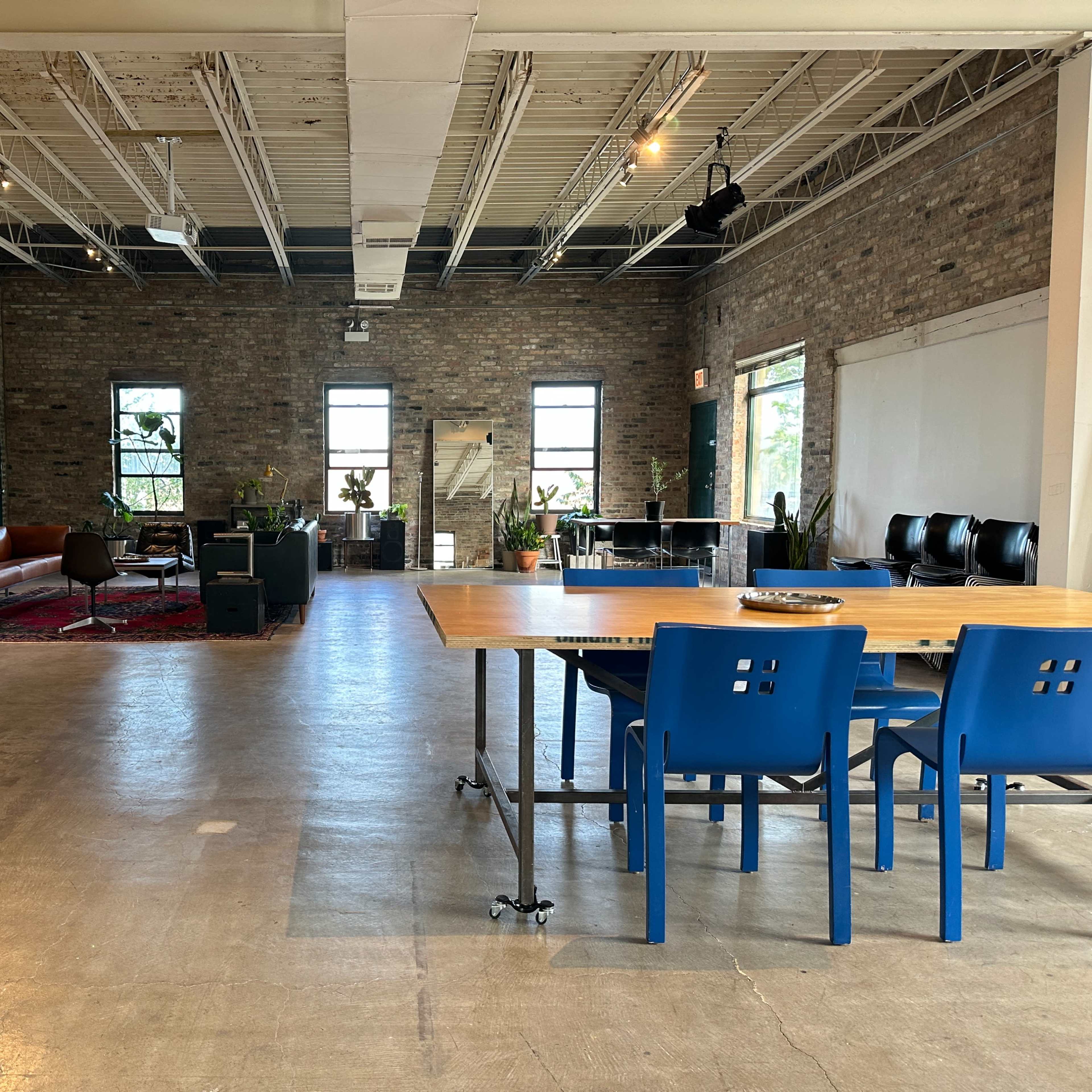 A spacious room with exposed brick walls, a wooden dining table with blue chairs, and a seating area featuring a sofa and plants.