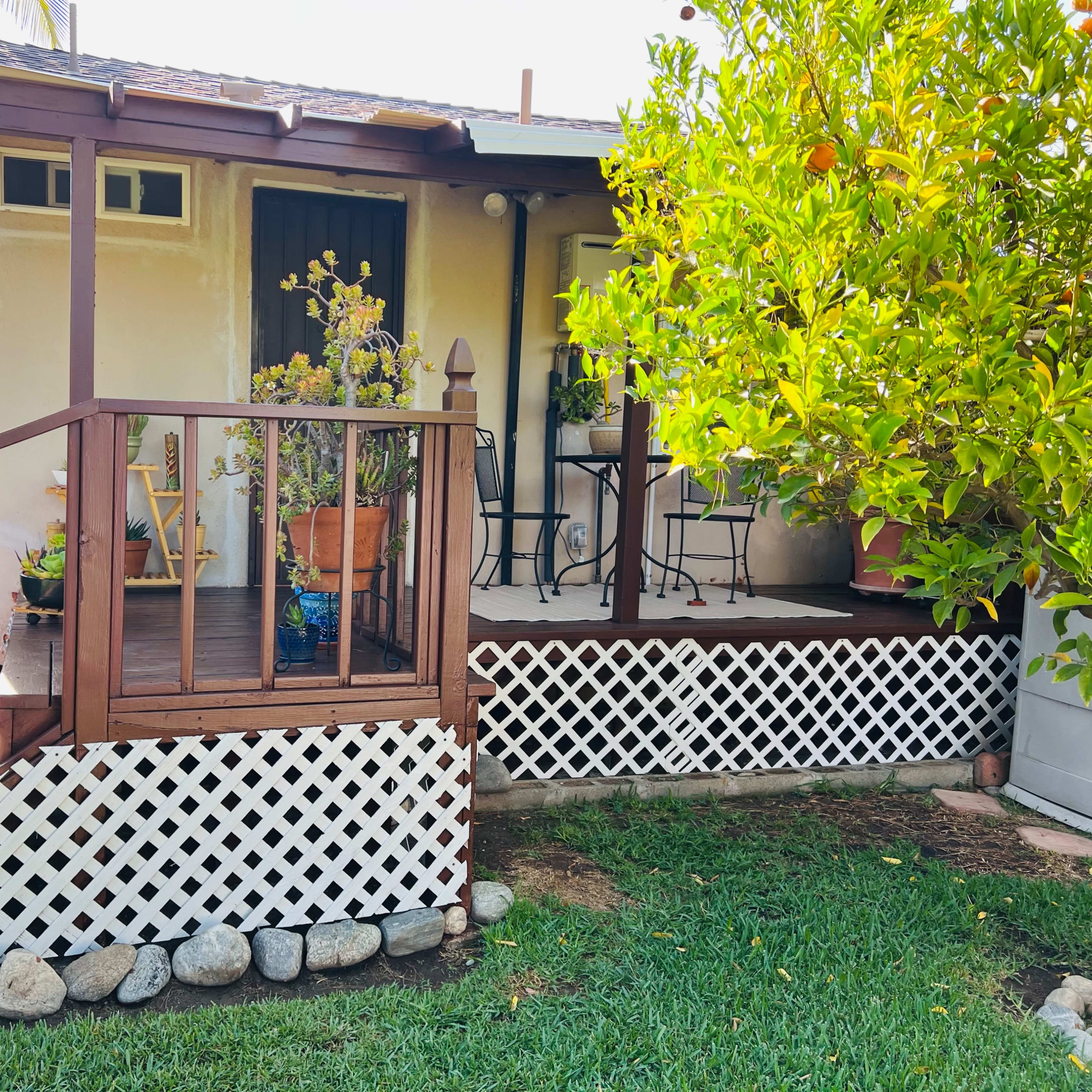 A small house features a wooden deck with a railing, potted plants, and chairs beside a garden with greenery.