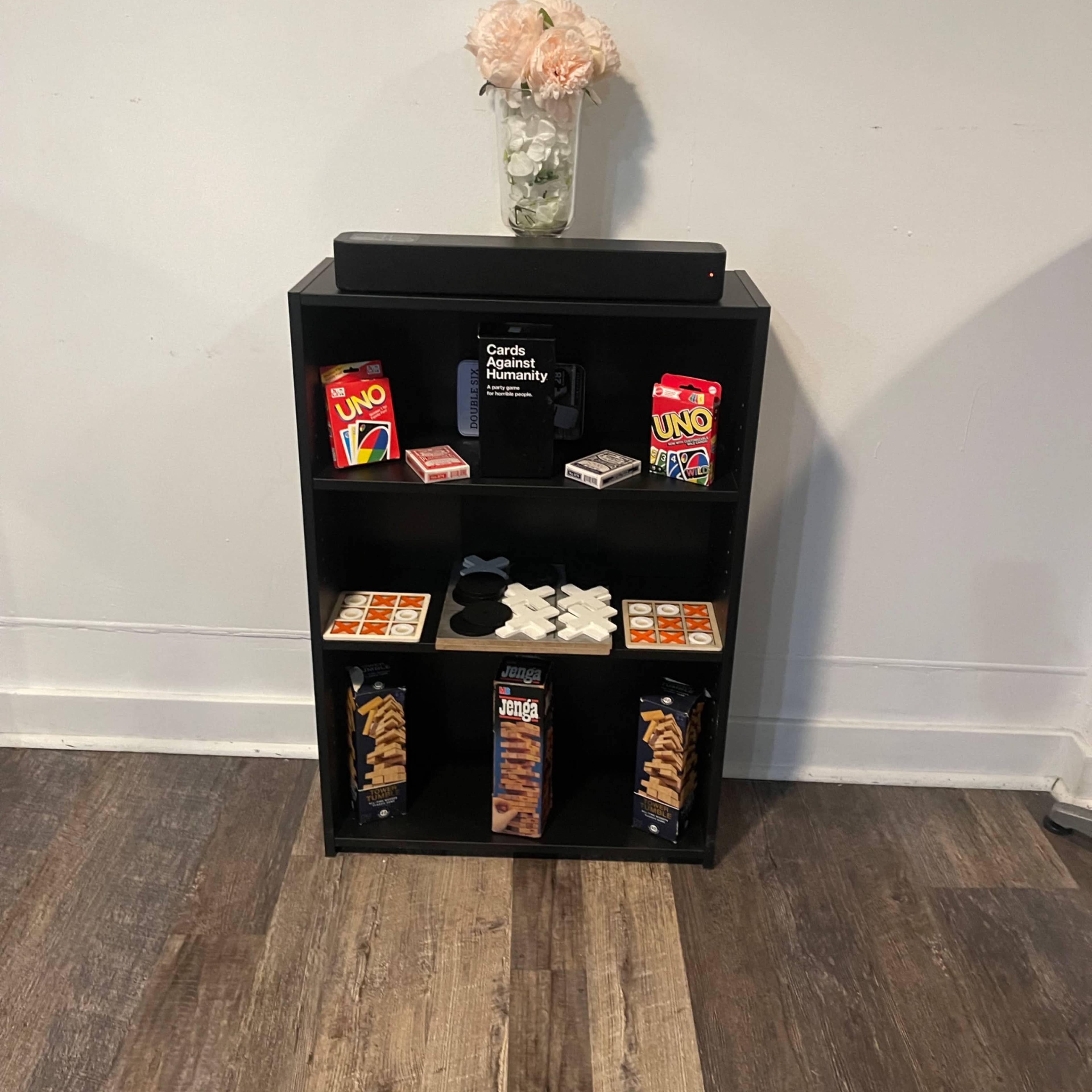 A black shelving unit displays several board games and a decorative flower arrangement on the top shelf.