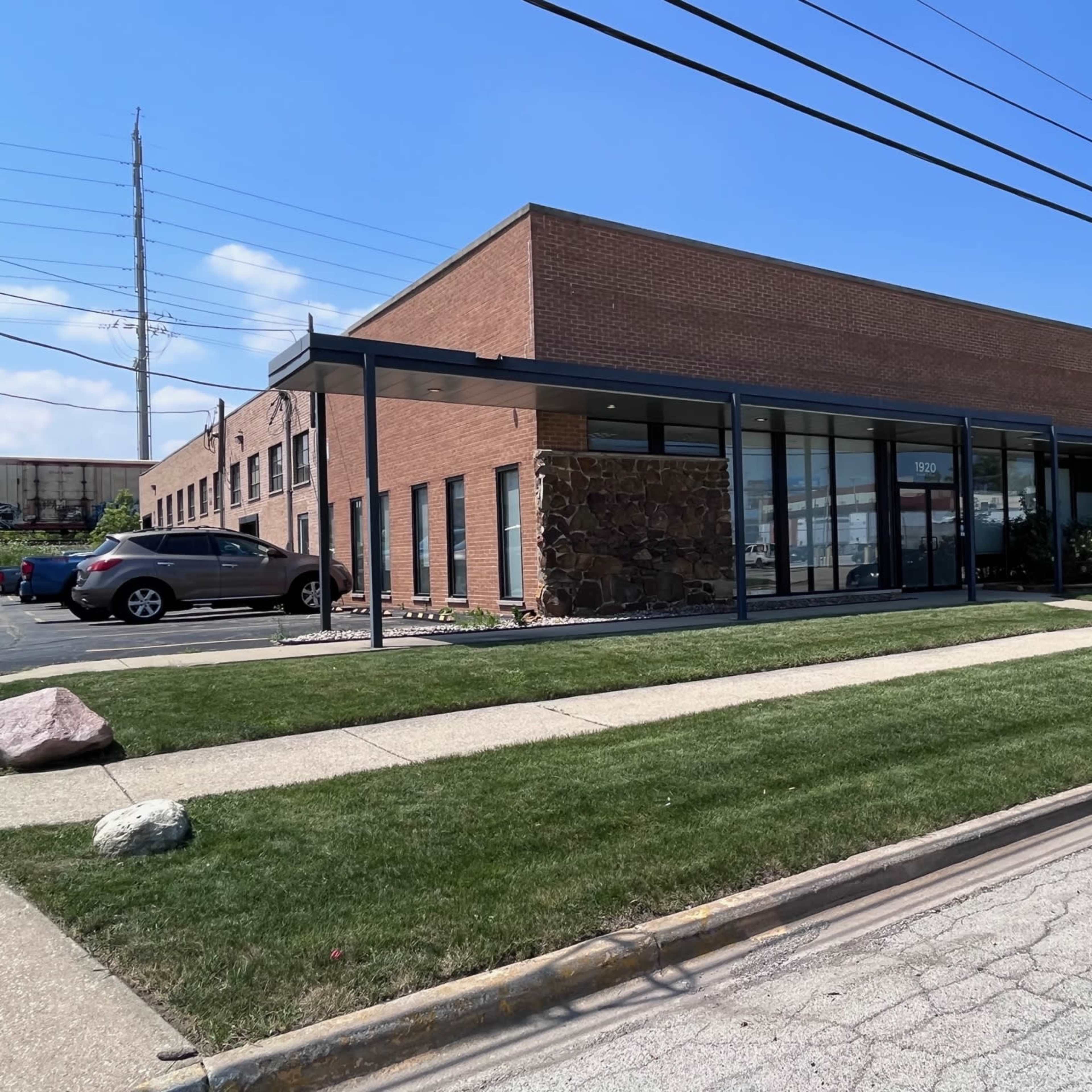 A single-story brick office building with large front windows and a landscaped area is located beside a paved parking lot.
