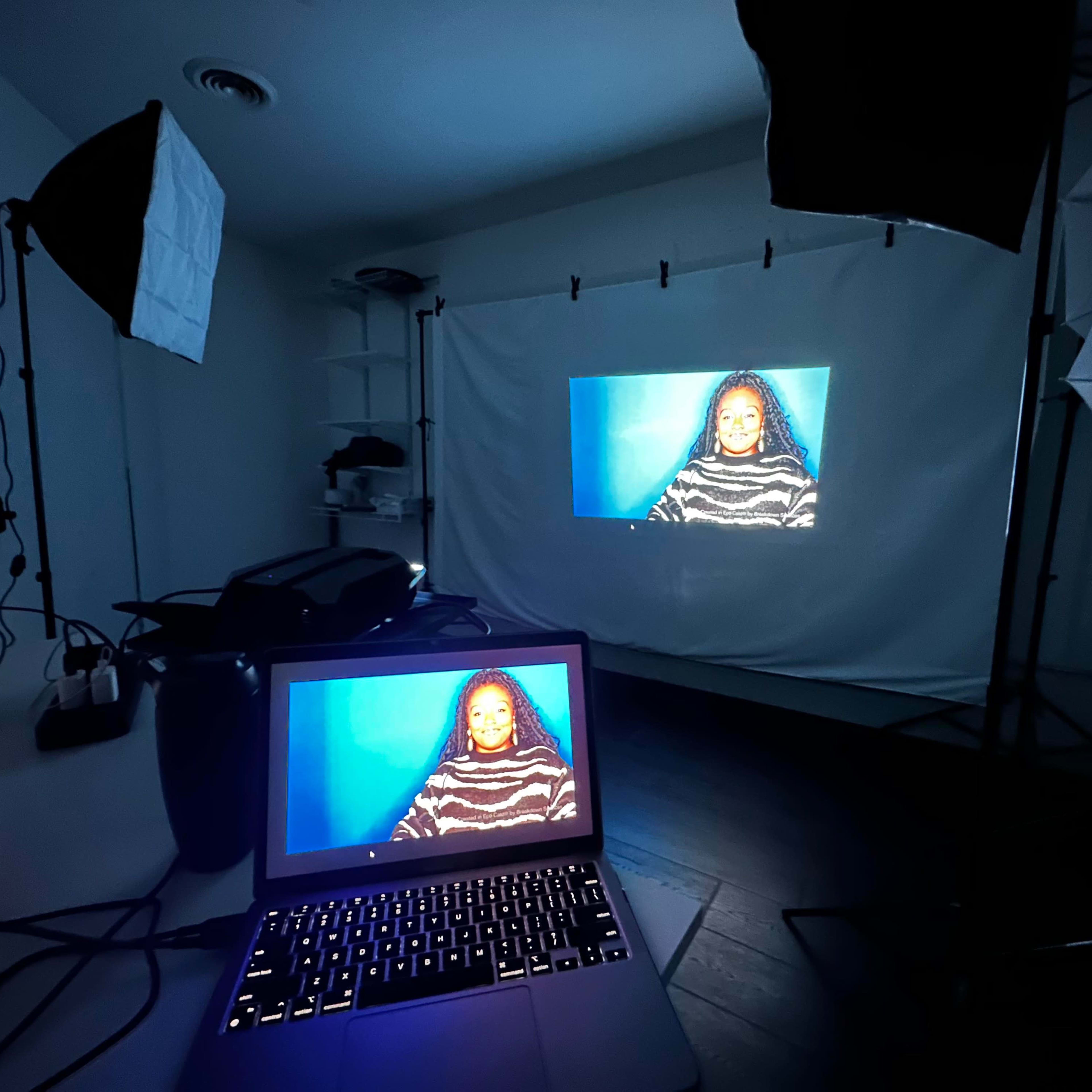 A computer displays an image of a person against a backdrop that is projected onto a white screen in a dimly lit studio.