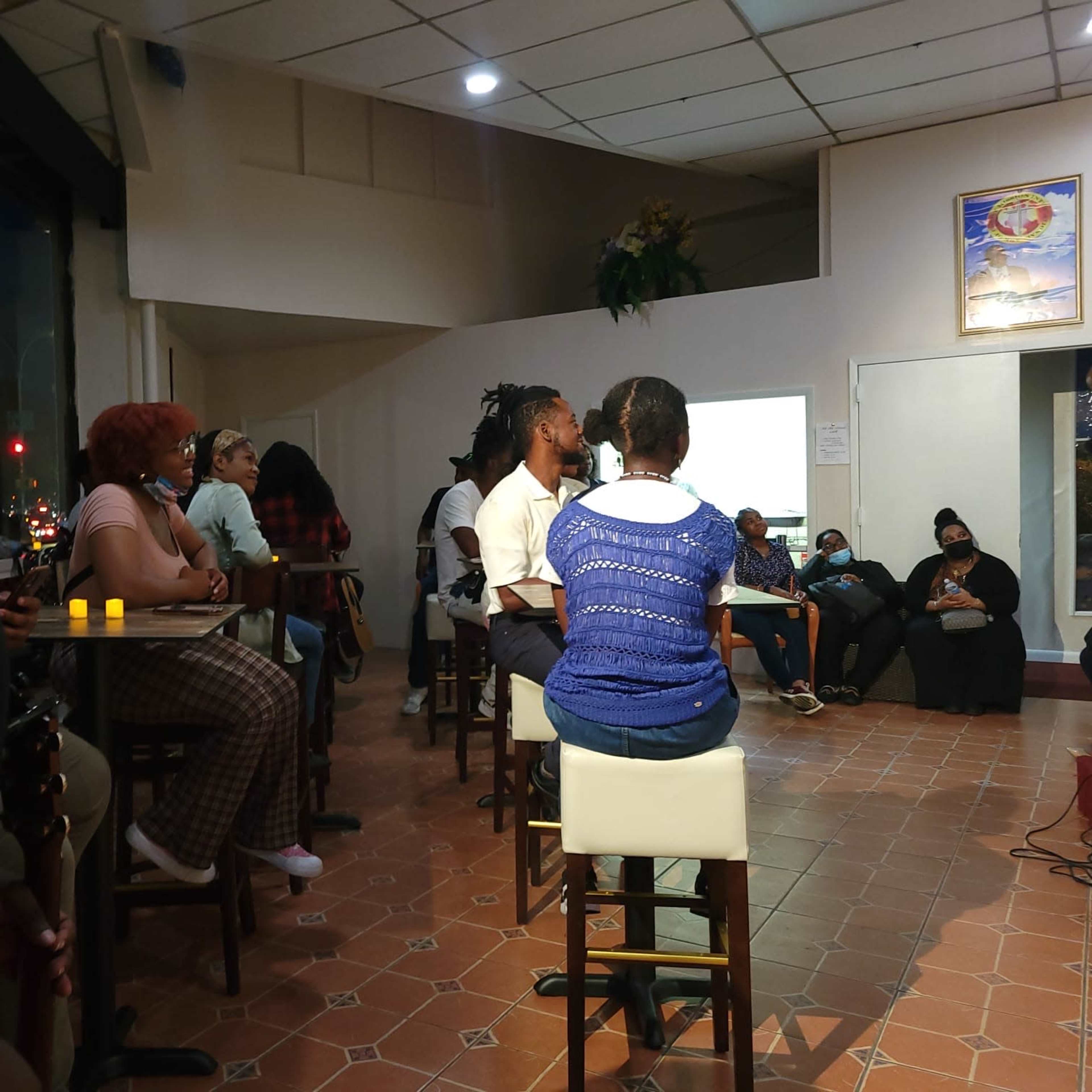 A small gathering of people seated at a bar and along the walls of a dimly lit room, with some individuals facing a speaker at the front.