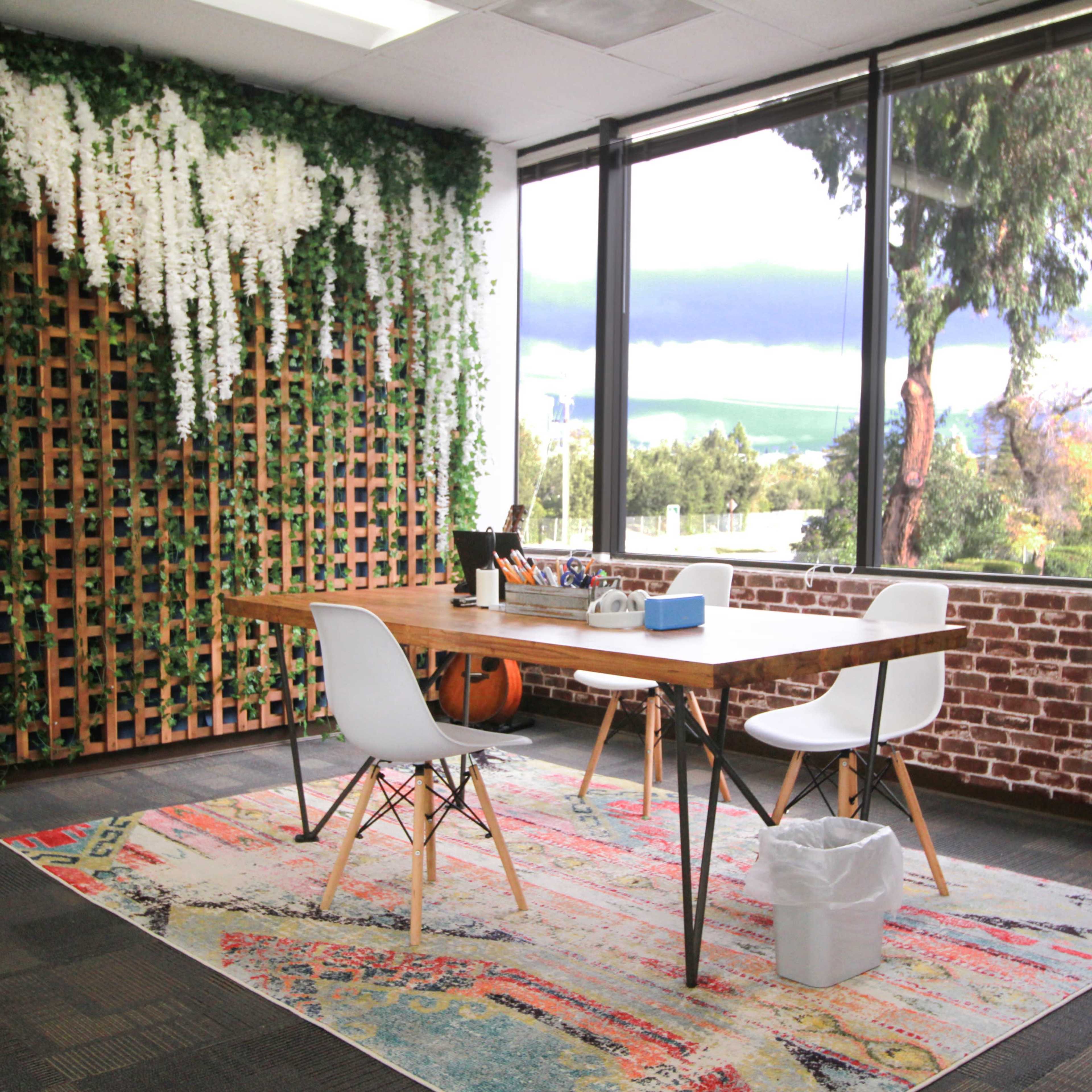 A modern office interior features a wooden table surrounded by white chairs, set against a backdrop of green vines and a large window showcasing palm trees outside.