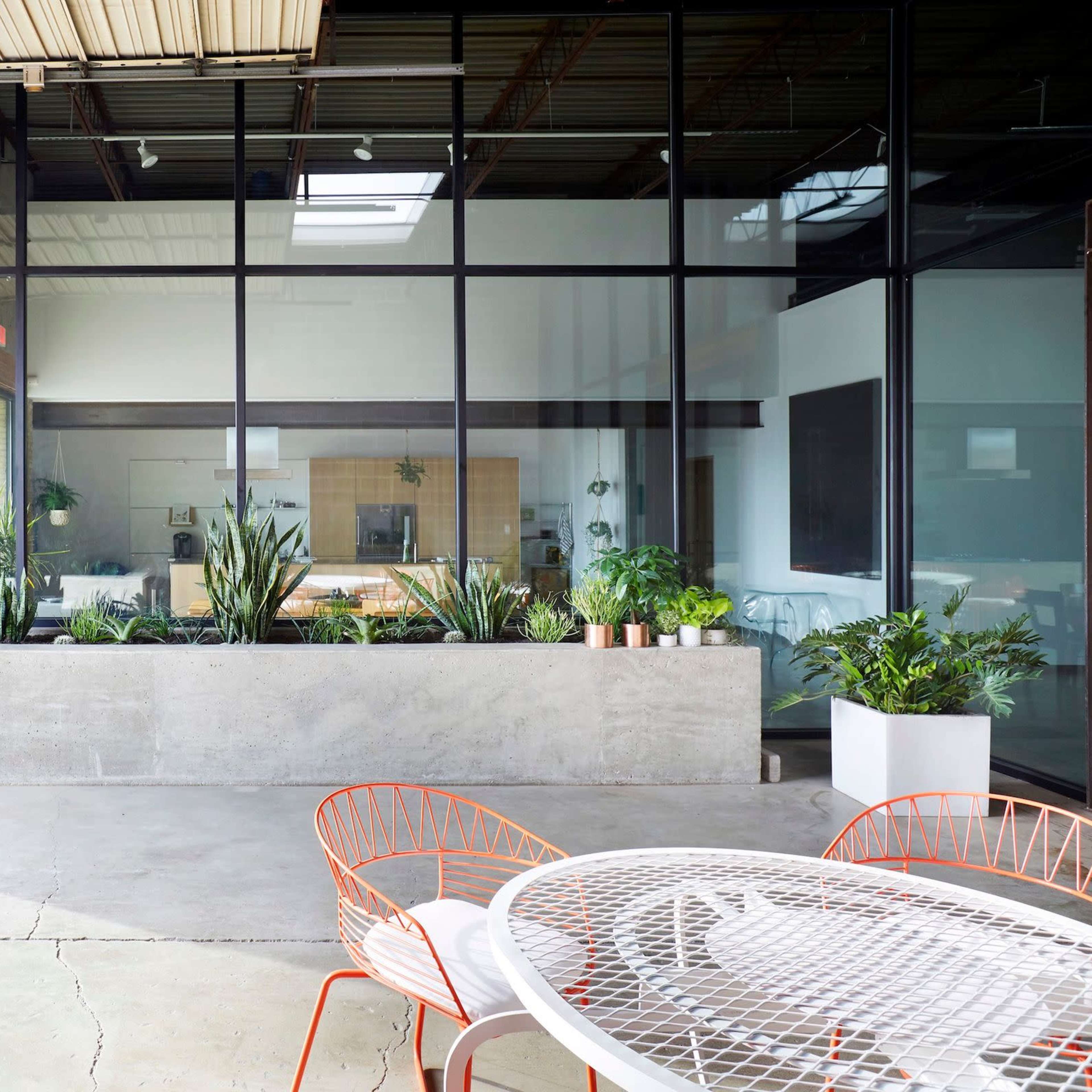 A modern indoor space with large glass windows, a concrete planter, and an outdoor seating area featuring a round table and orange chairs.