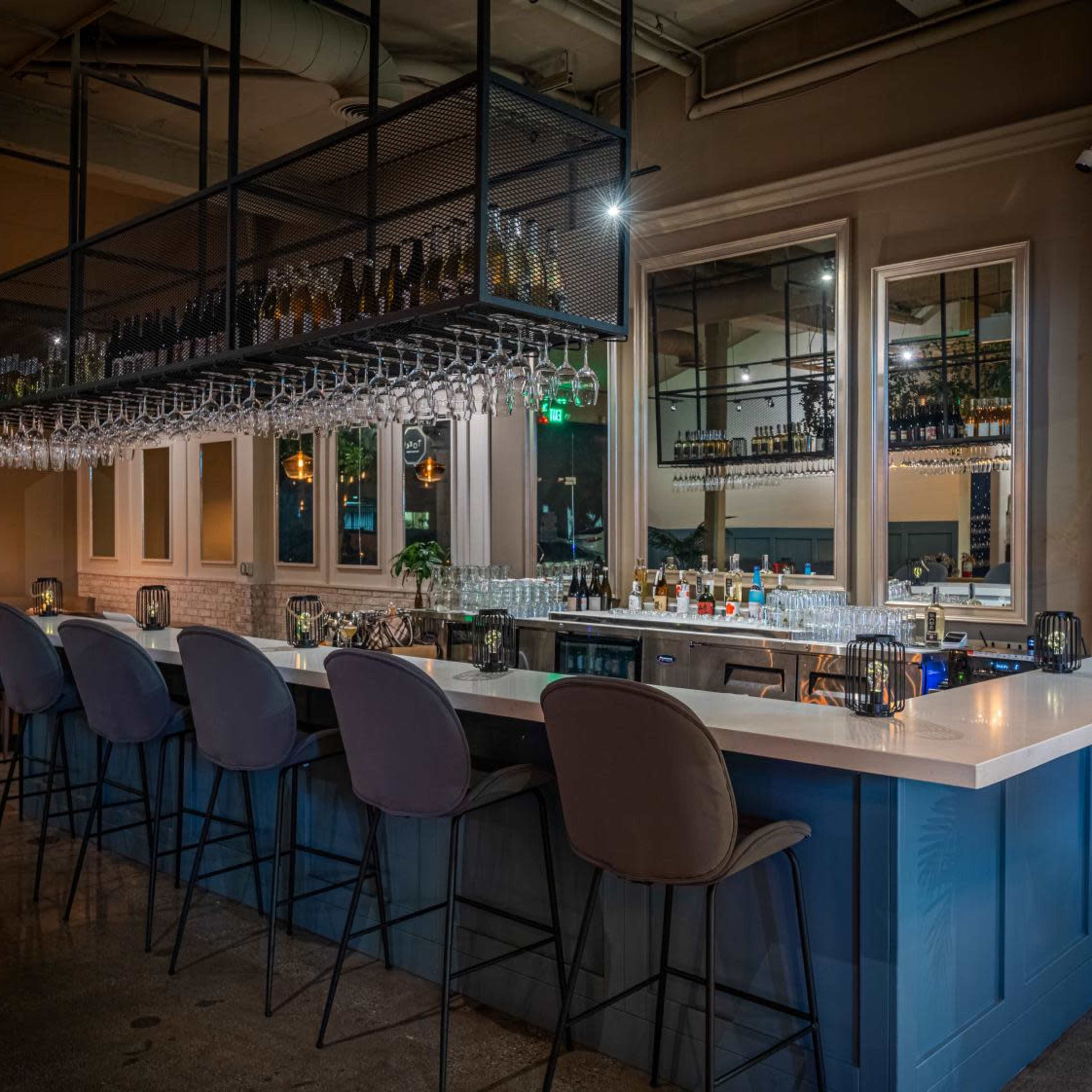 A modern bar area with a white countertop, bar stools, and a rack of wine bottles overhead.