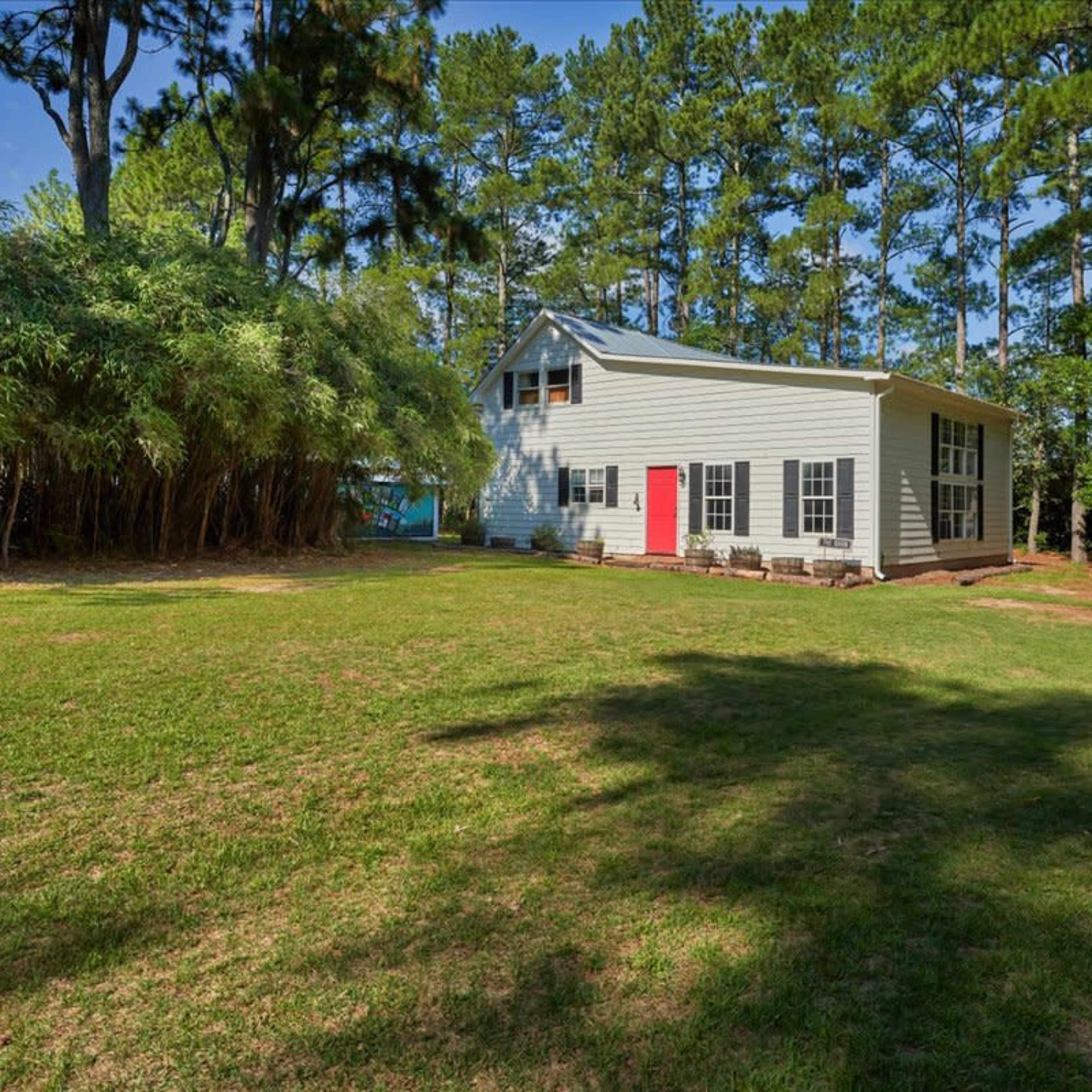 A two-story gray house with a red door is set in a grassy yard surrounded by tall trees and bamboo.