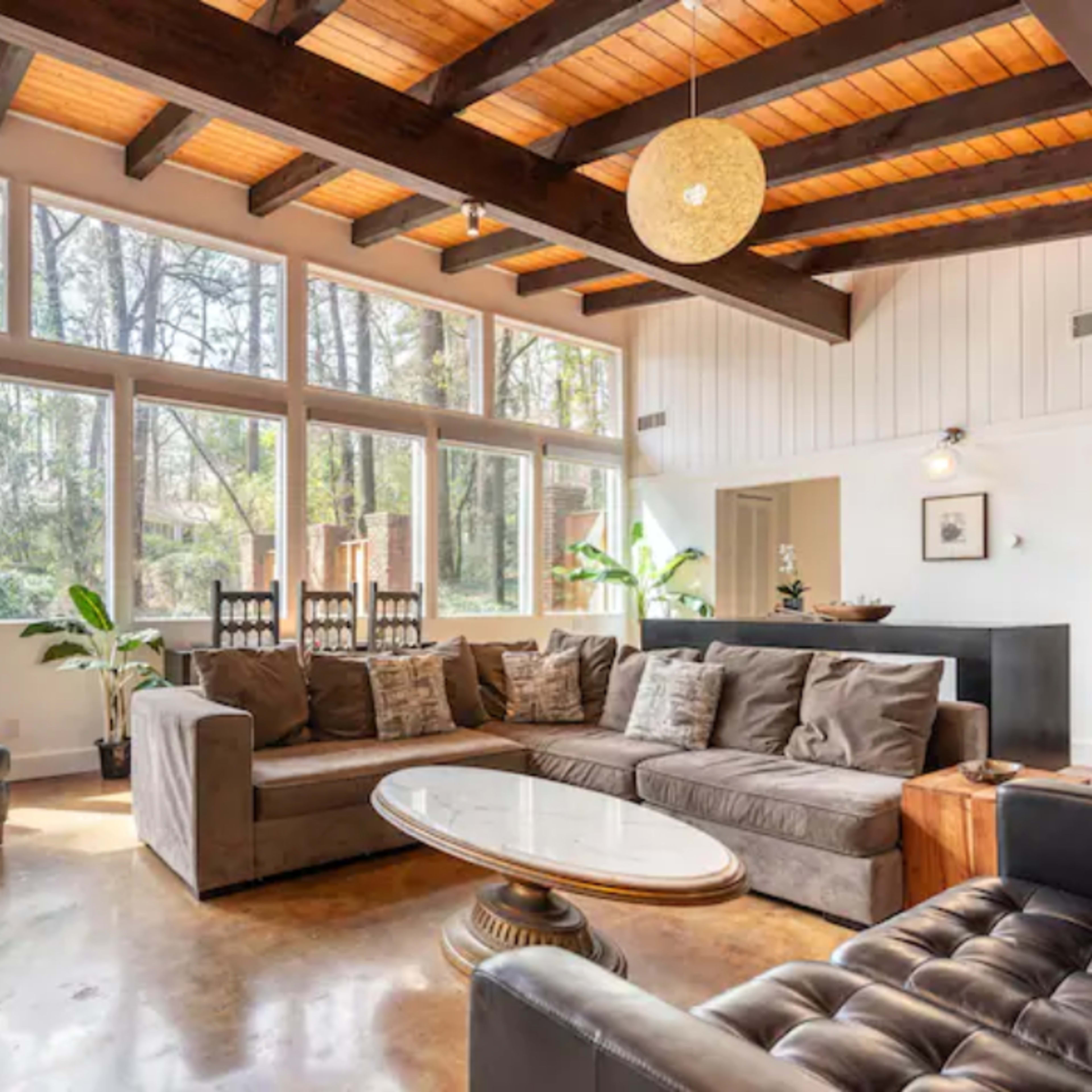 A modern living room with large windows allowing natural light, featuring a sectional sofa, a round coffee table, and wooden beams on the ceiling.