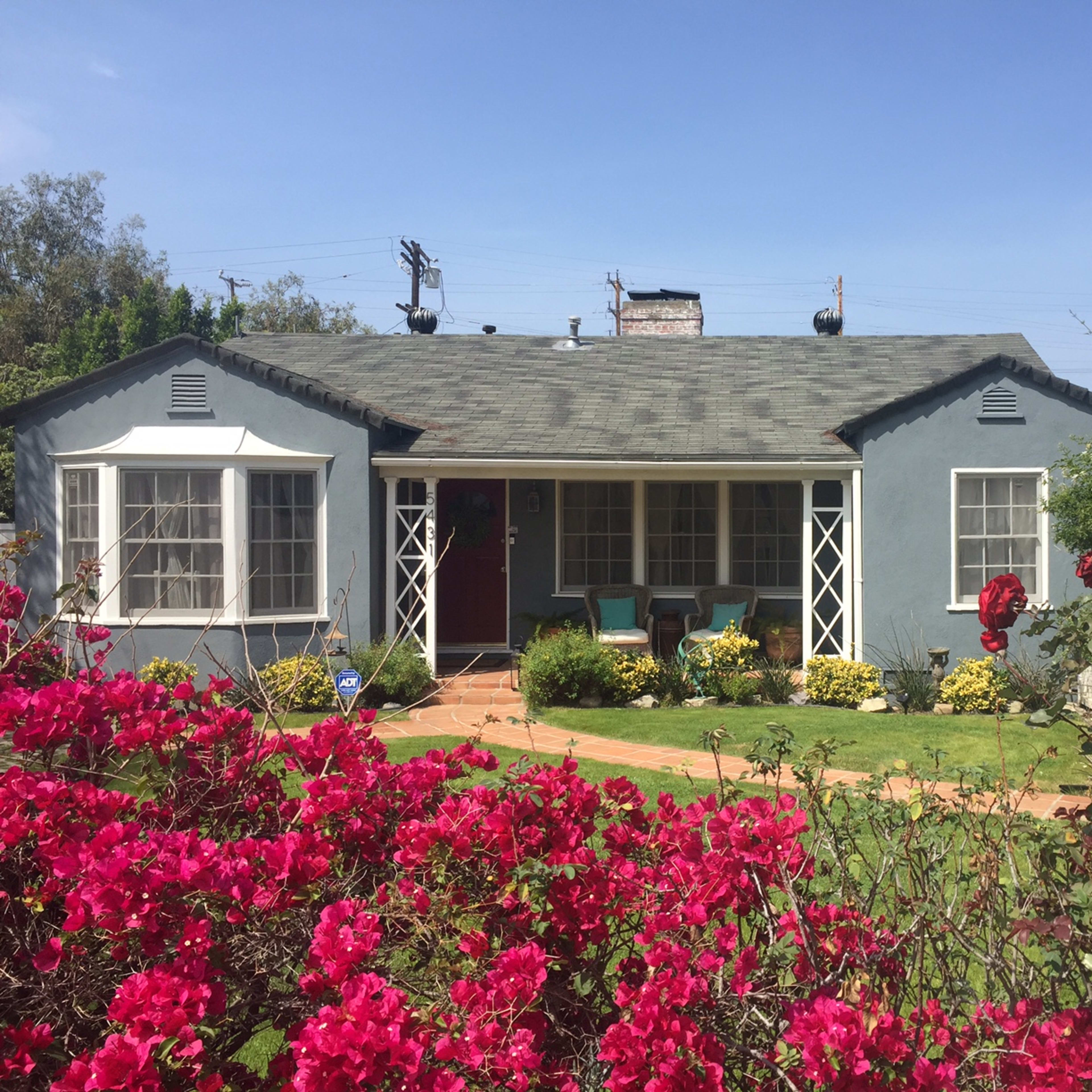 A light blue house with a shingled roof is surrounded by colorful flowers and features a red front door along with a small porch.