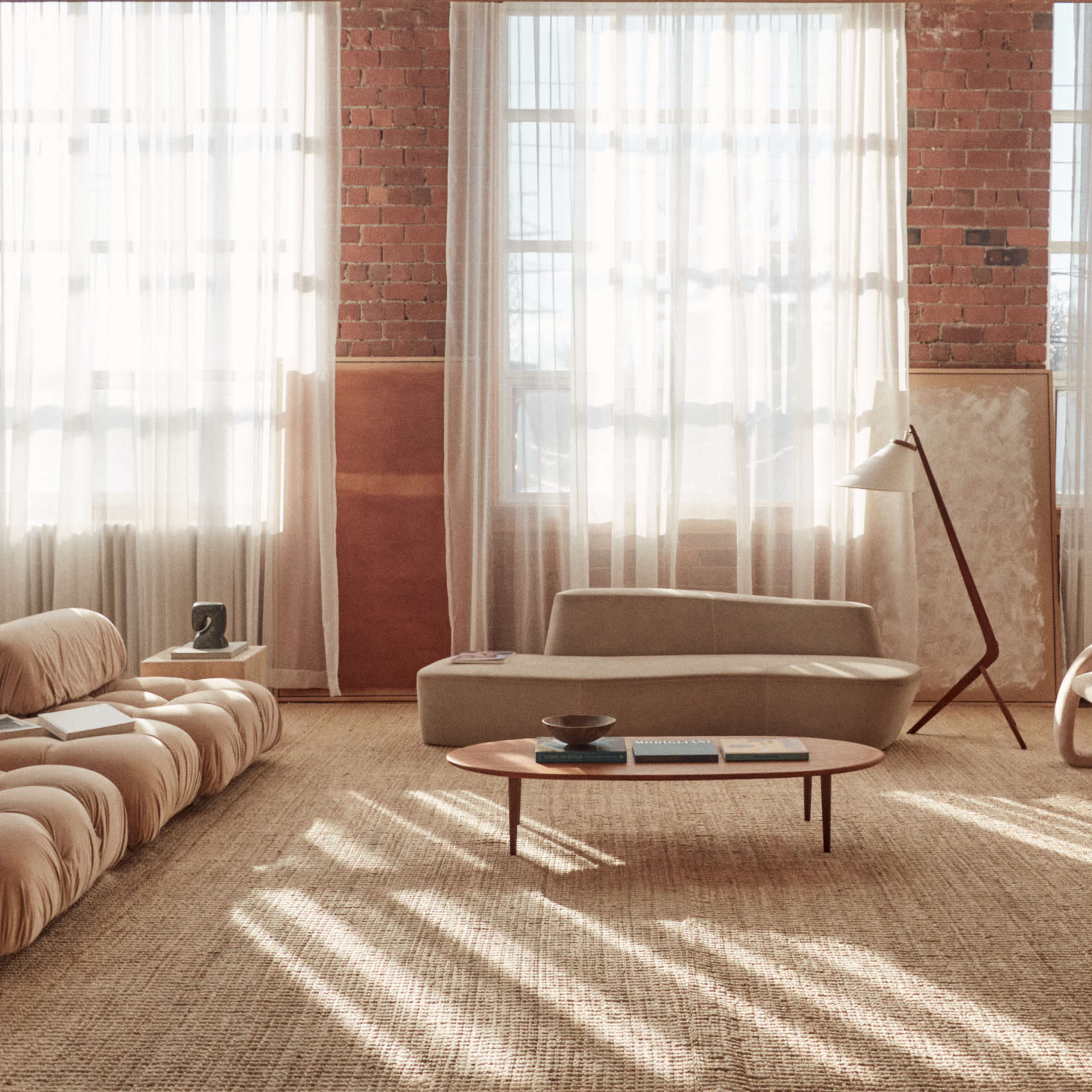 The scene features a spacious living room with large windows, showcasing minimalist furniture including a beige sofa, a curved chair, and a wooden coffee table on a textured rug.